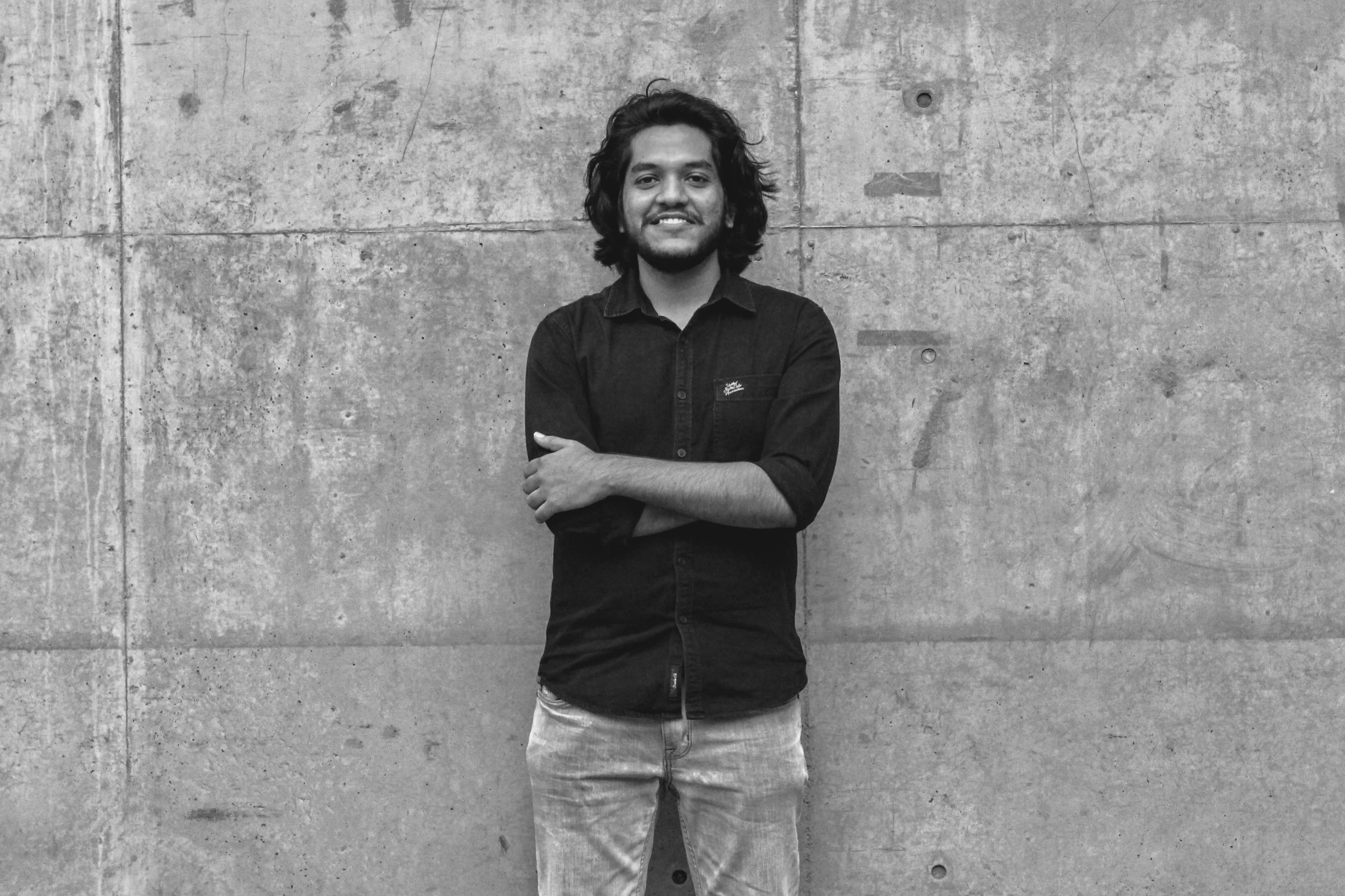 Modern architectural workspace with large floor-to-ceiling windows, surrounded by trees, and desks filled with design materials and books. Two blurred figures are walking through the space.