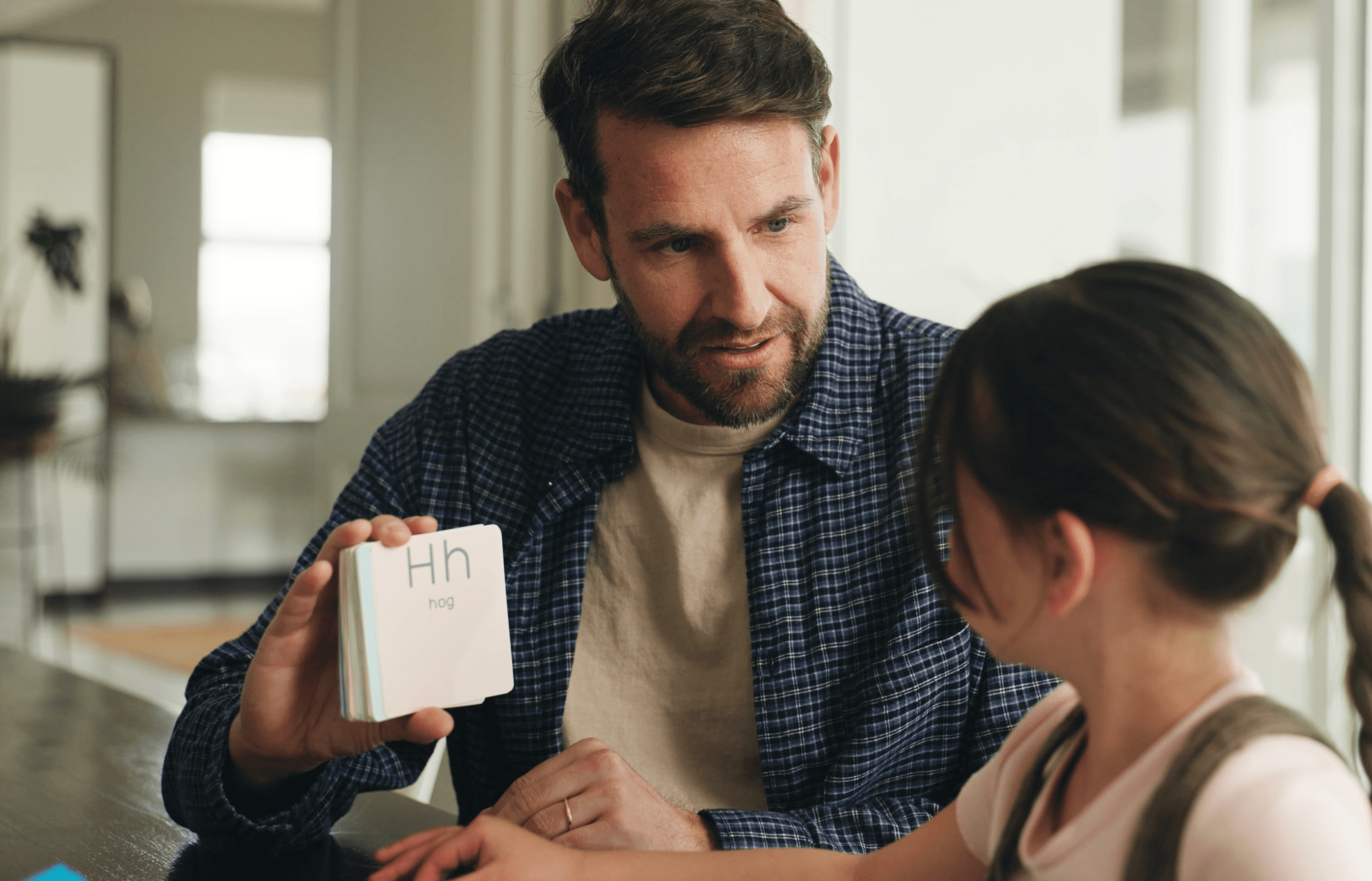 Man with glasses and tied-back hair writing on a tablet while seated at a desk in a softly lit room.