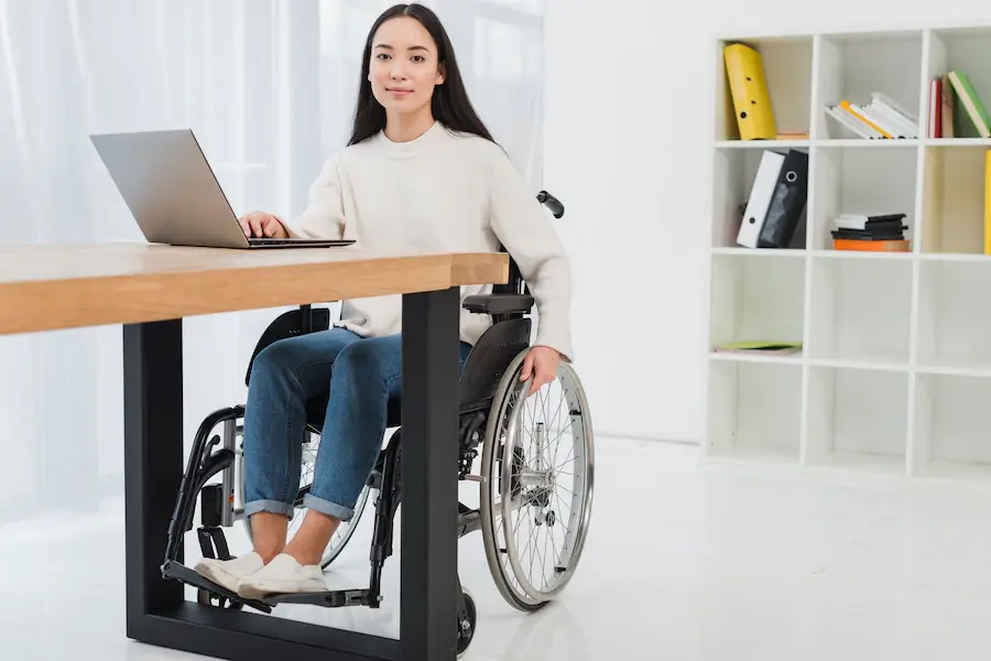 A imagem mostra uma jovem mulher em uma cadeira de rodas, trabalhando em um laptop que está sobre uma mesa de madeira. Ela veste um suéter bege e calça jeans, com uma expressão tranquila e confiante. O ambiente ao seu redor é claro e organizado, com uma estante ao fundo contendo arquivos e livros. A cena transmite uma sensação de profissionalismo e inclusão, mostrando uma mulher em um espaço de trabalho acessível, envolvida em suas atividades com independência e foco.