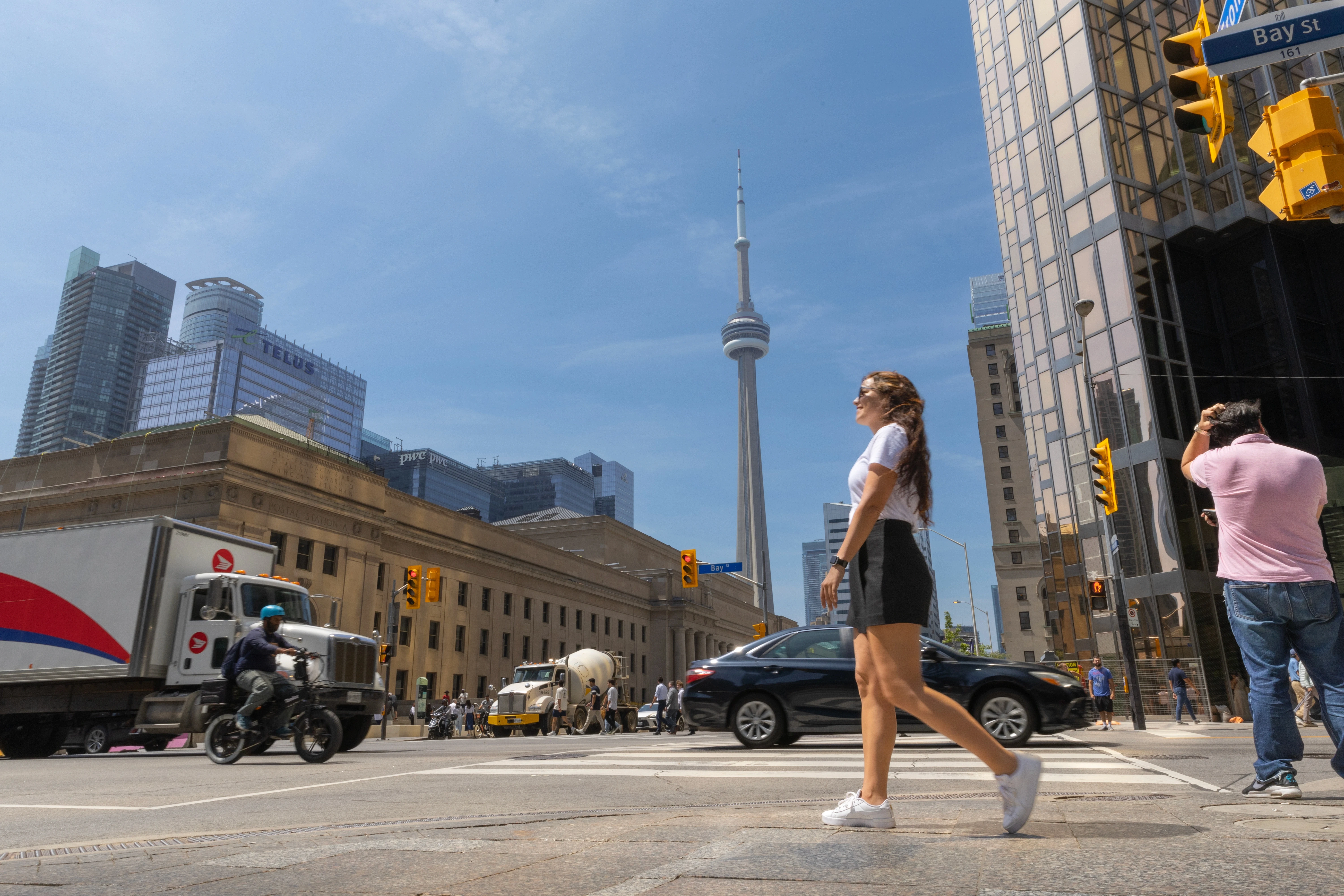 Mulher caminhando no centro de Toronto em um cruzamento com o CN Tower ao fundo, em dia ensolarado.
