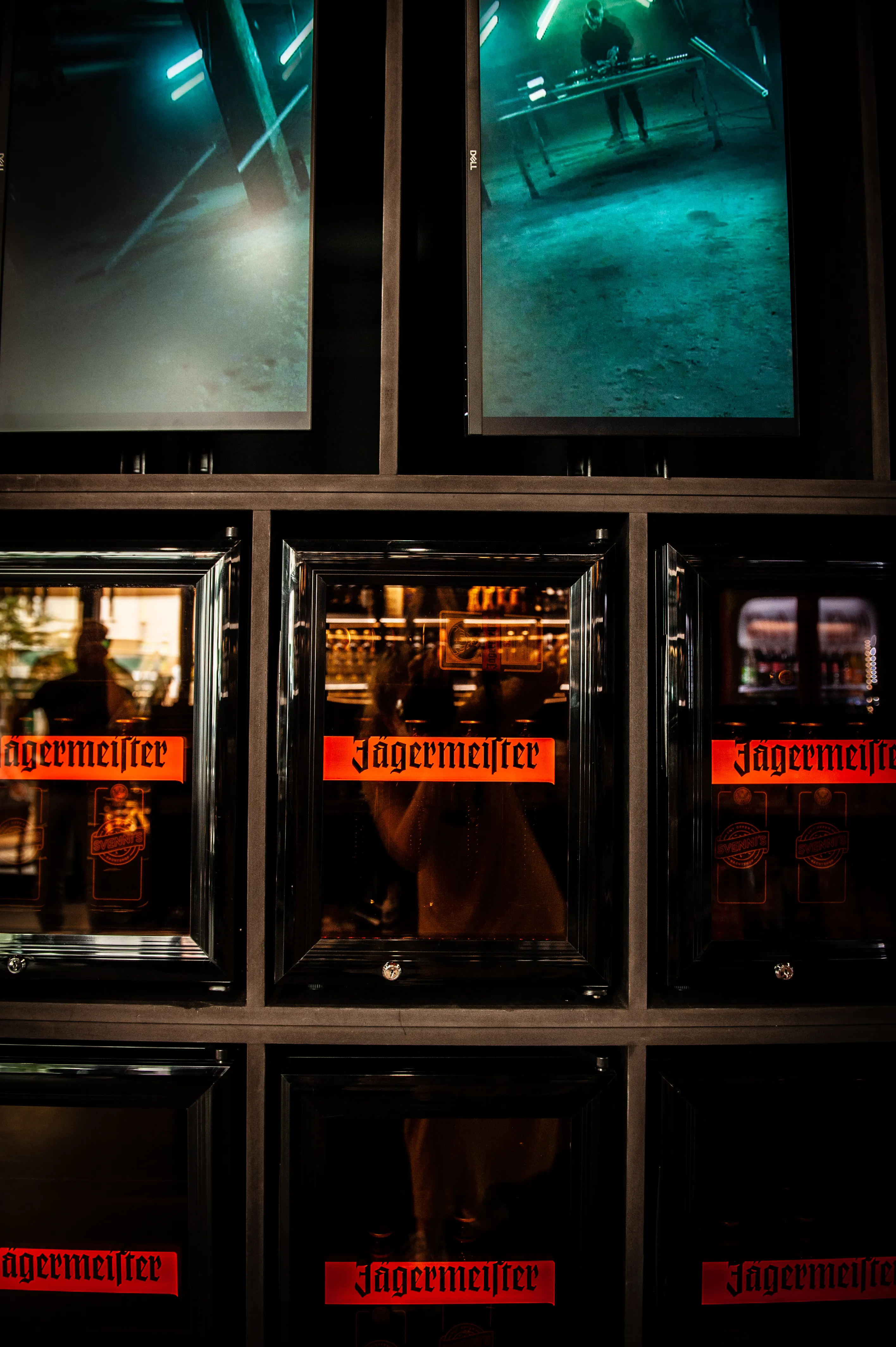 Grid of Jägermeister-branded fridge doors with mirrored fronts at JÄGERMEISTER x SVENNI'S, topped by screens showing teal-lit club scenes.
