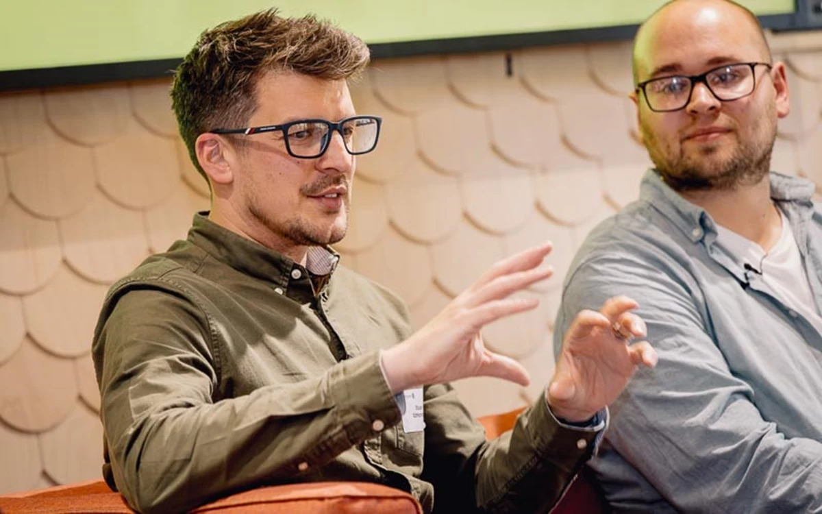 Two panelists sit in discussion, with one gesturing while explaining a point. The setting suggests an informal talk or panel at a professional event.
