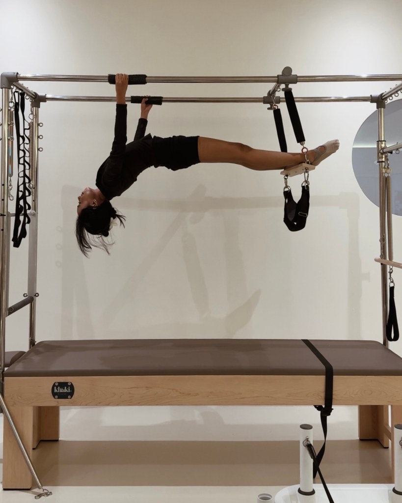 A person performing an advanced exercise on a Pilates reformer, hanging upside down from a horizontal bar, with legs extended and supported by straps.