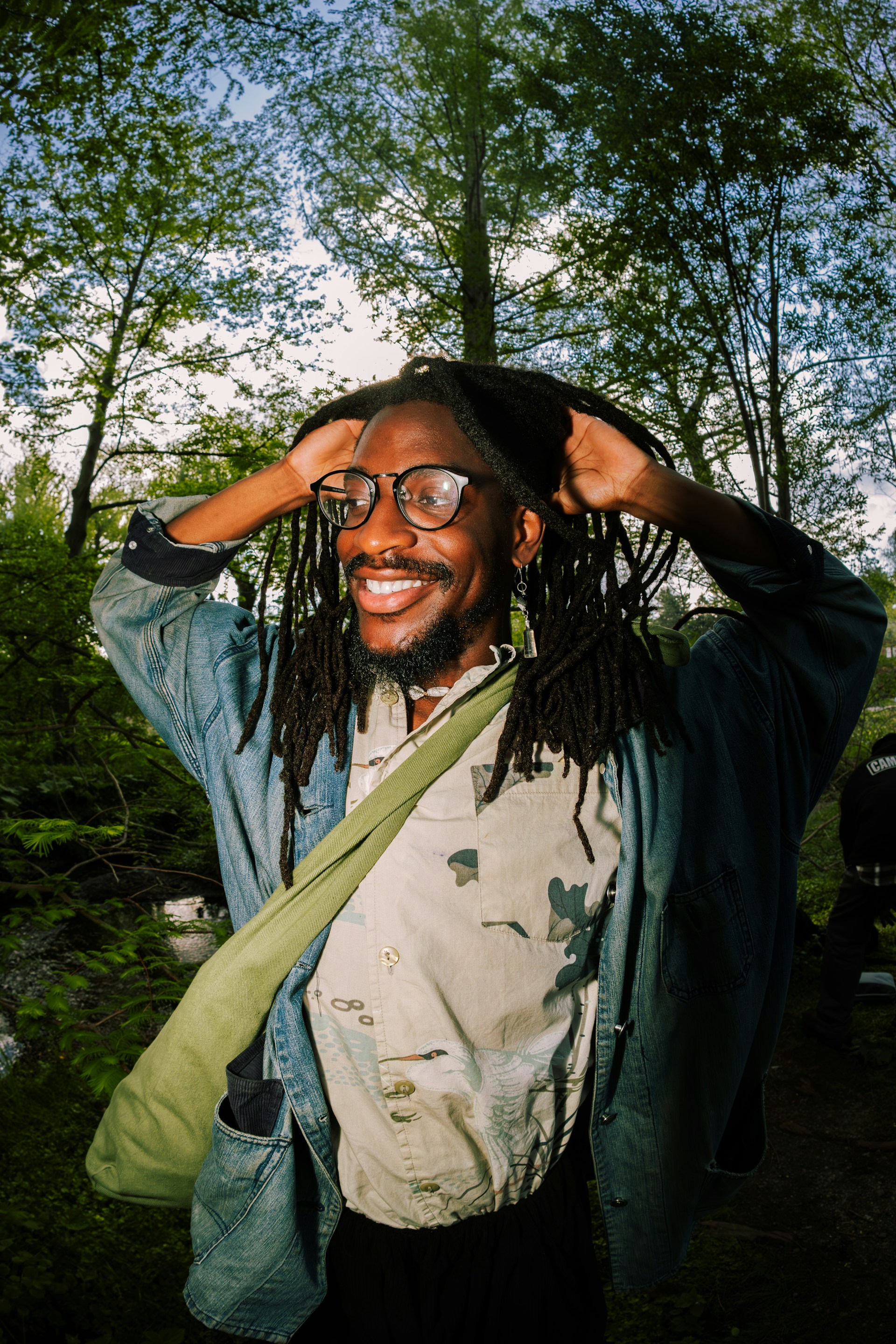 Smiling man with dreadlocks and glasses standing in a forest.