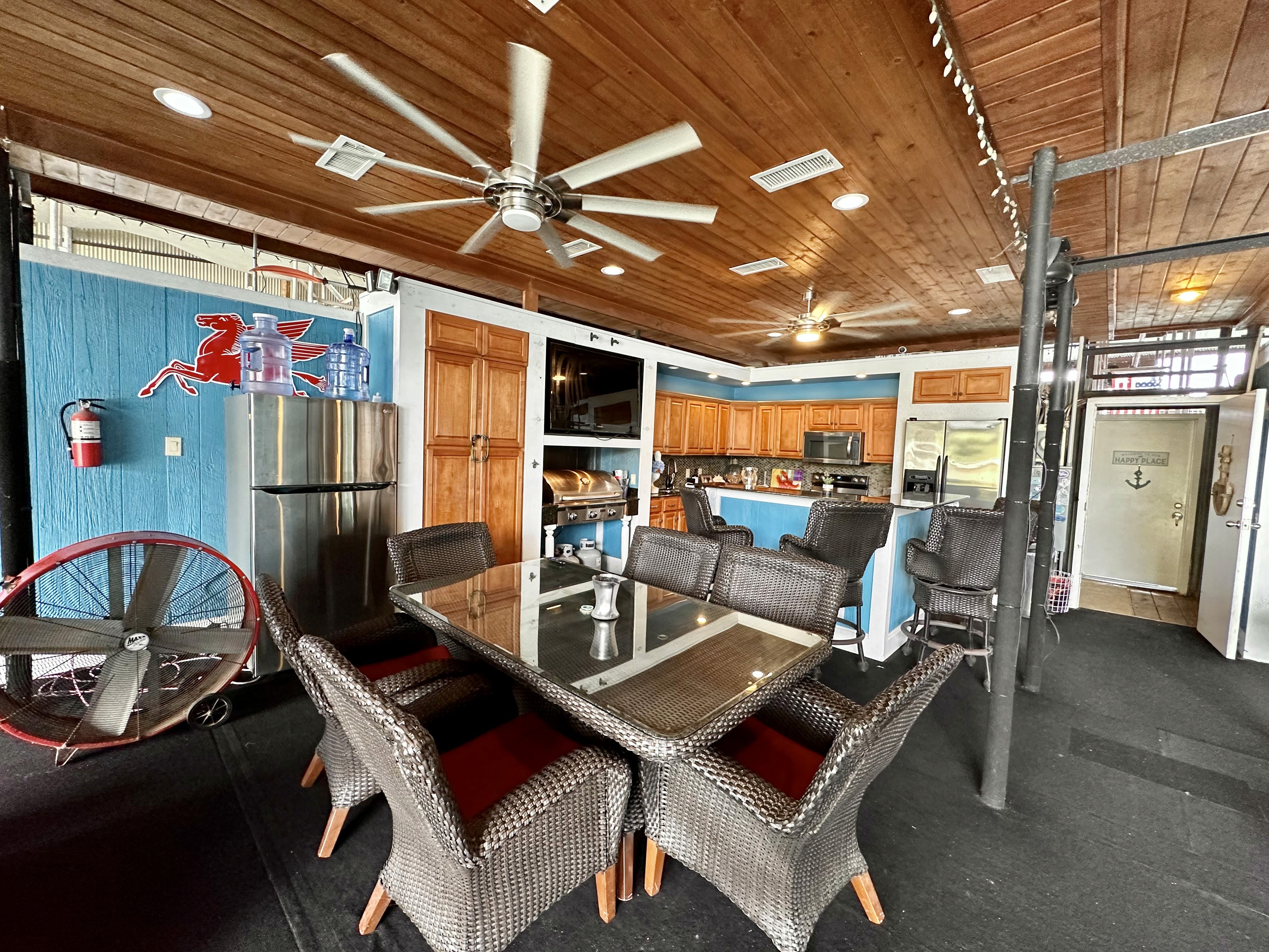 A rustic indoor patio features a wicker dining set with a glass table, ceiling fans, wooden ceiling, and a bar area with stainless steel appliances, against a backdrop of blue and wooden paneled walls.
