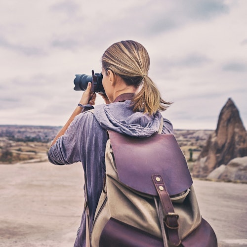 一位身穿连帽衫并携带大背包的人正在岩石遍布的户外景观中用相机拍照。