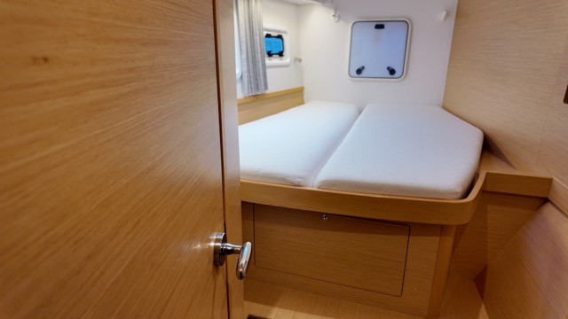 Lagoon 42 catamaran forward hull double cabin viewed through the open light oak veneer cabin door, showing a wide double berth with white fitted mattress, under-berth storage drawers, hull portlight window, and a wall-mounted tablet screen on the cabin bulkhead