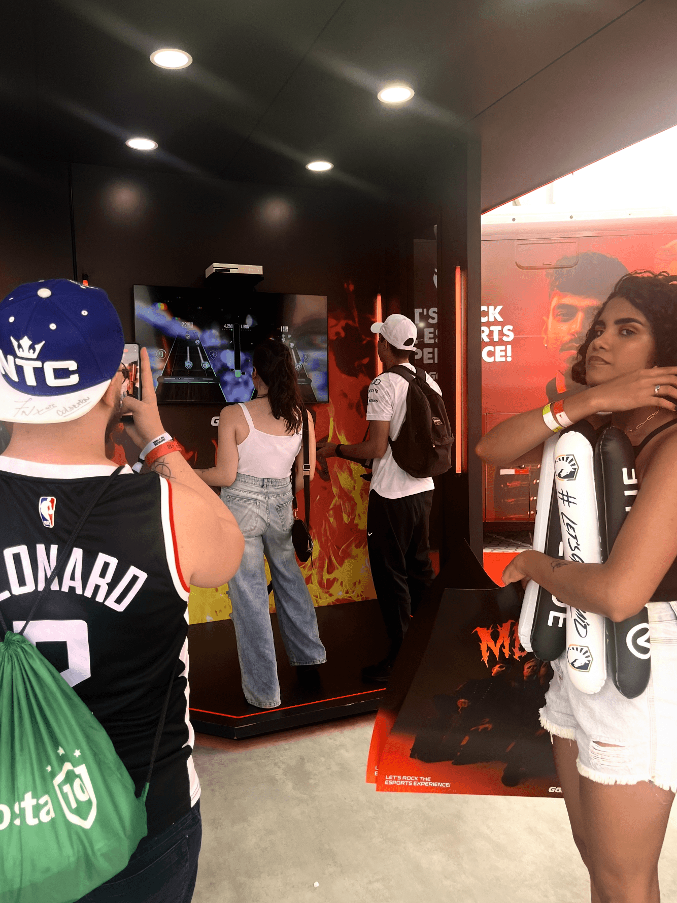 Visuel du projet Metality : Un homme prend une photo de deux personnes jouant à un jeu de simulation de guitare, tandis qu'une femme à droite tient des affiches et des goodies de l'équipe Metality.