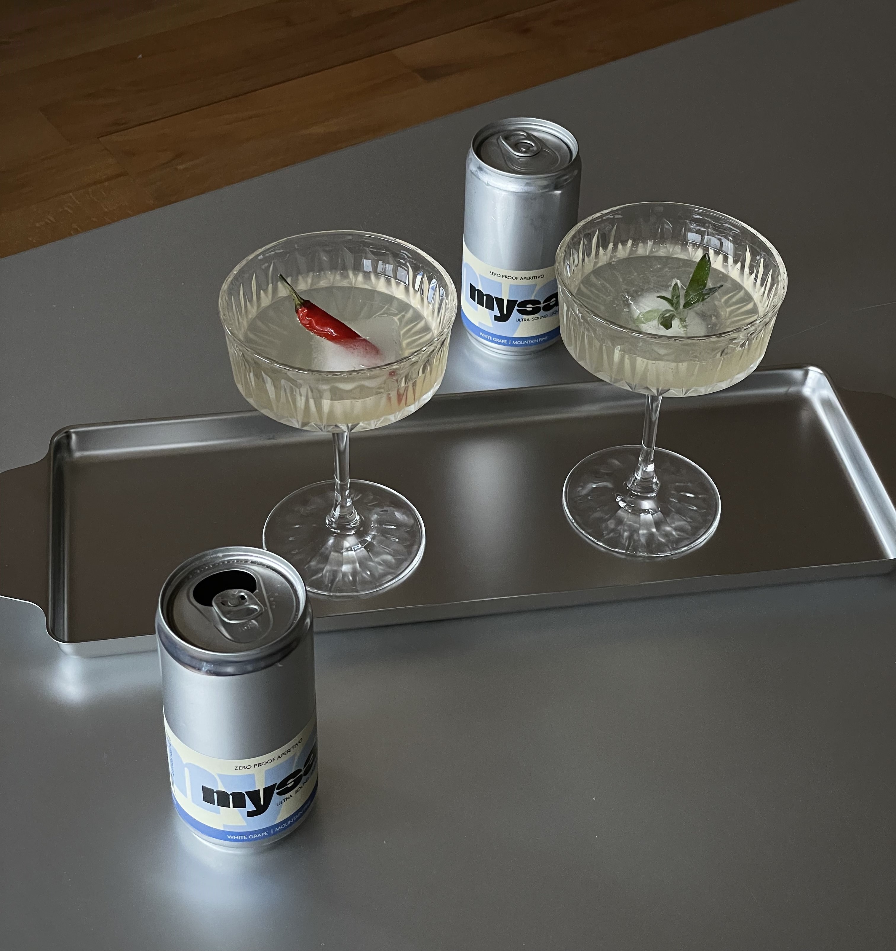 mysa poured into coupe glasses with chili and sage garnish on a metal tray, next to two cans