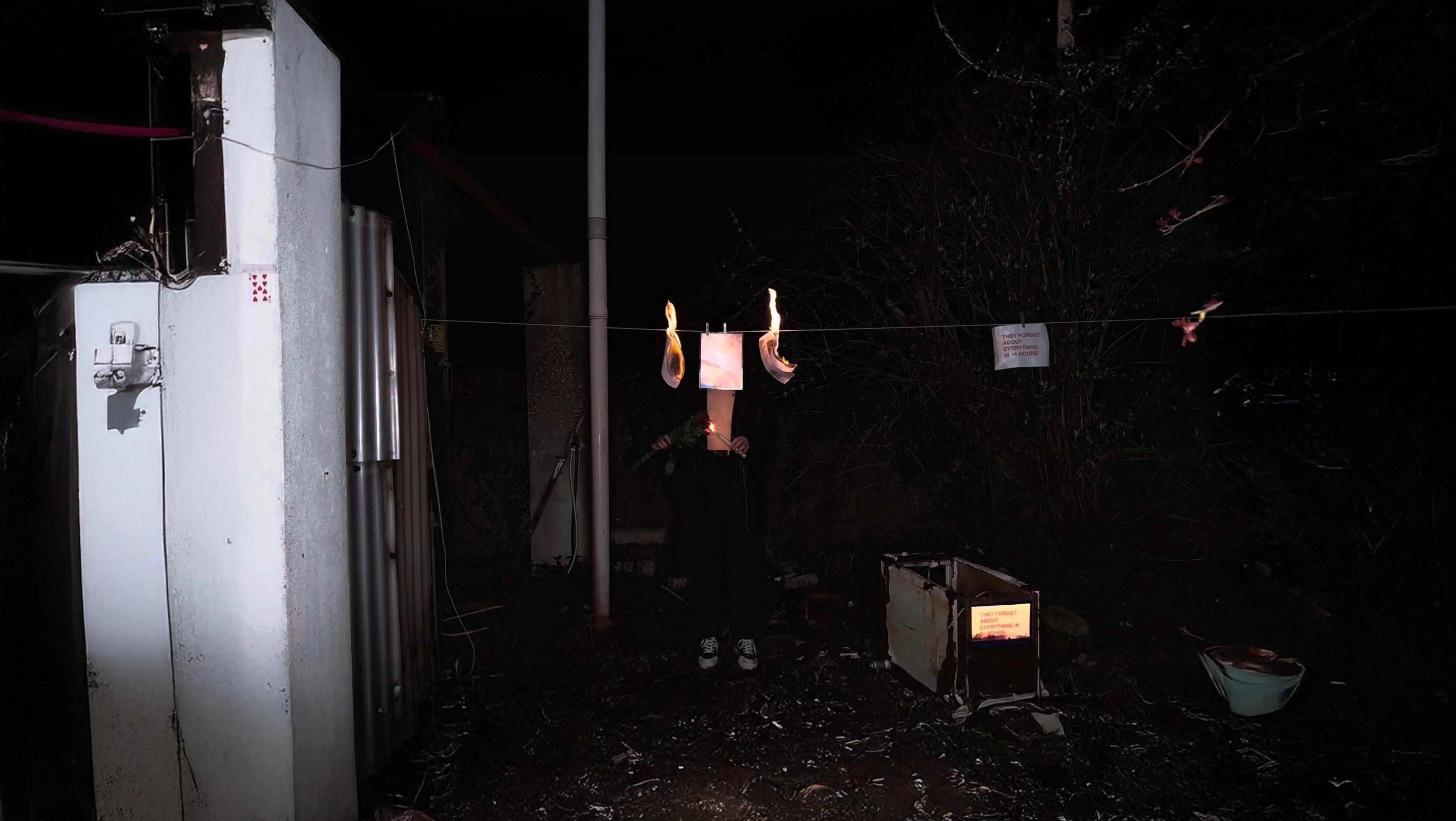 Dark nighttime scene of a figure standing behind burning papers clipped to a clothesline, surrounded by scattered debris, handwritten notes, and a glowing box. The dramatic lighting, flames, and shadows evoke themes of memory, erasure, protest, and impermanence.