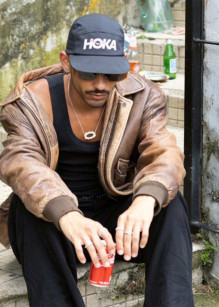 Man wearing a Hoka cap sitting on steps with a can of coca cola wearing silver jewelry