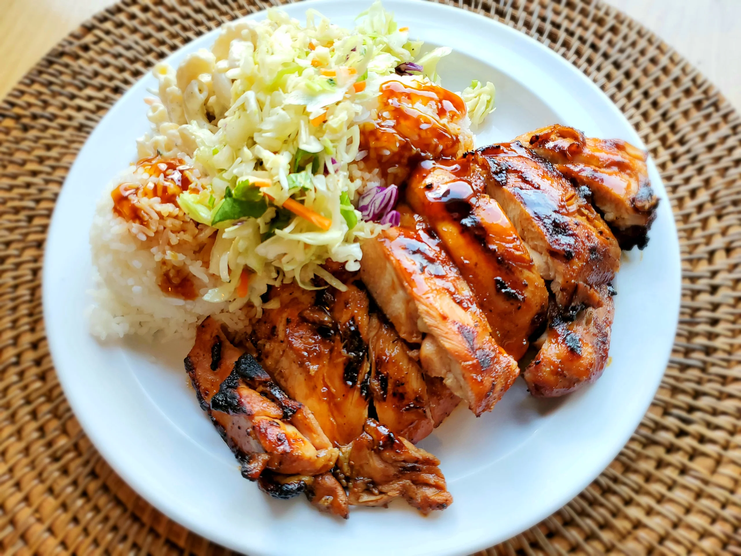 A white plate featuring sliced, char-grilled Huli Huli chicken thighs with a dark glaze, two scoops of rice, creamy mac salad, and a fresh vegetable slaw.