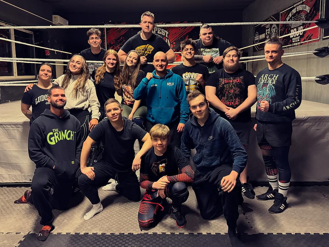 A group of six people gather around a wooden table where an older man in an apron kneads dough