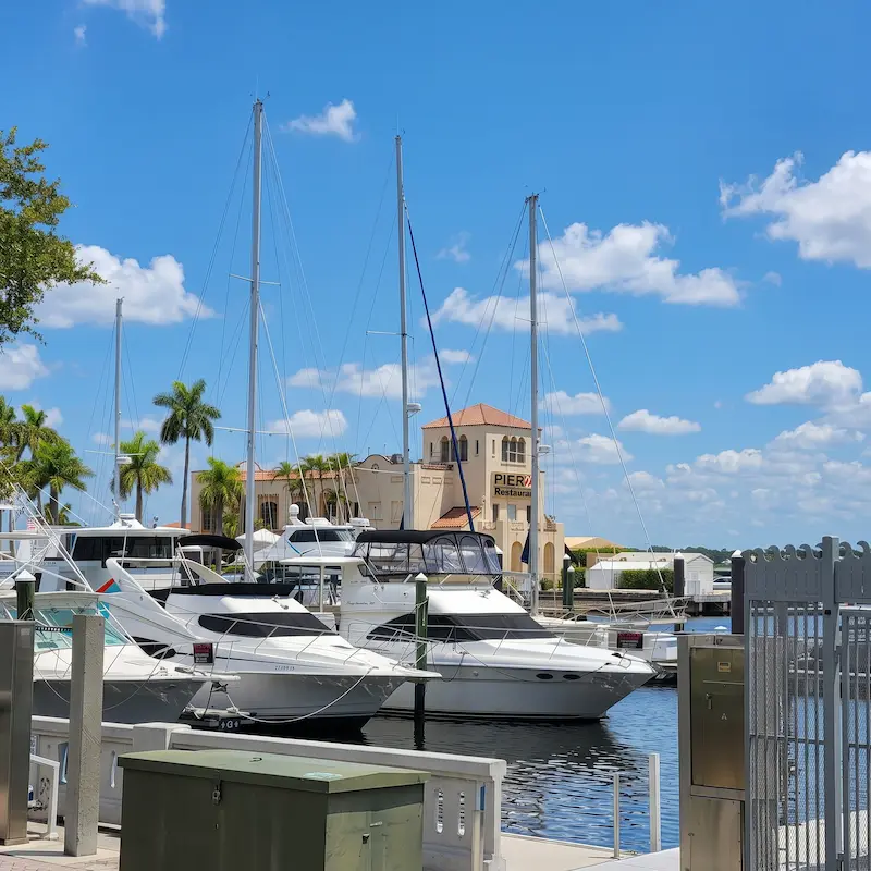 Scenic view of Bradenton Riverwalk, FL, featuring a marina with several moored yachts against a backdrop of palm trees and a building marked "Pier 22" under a partly cloudy sky.