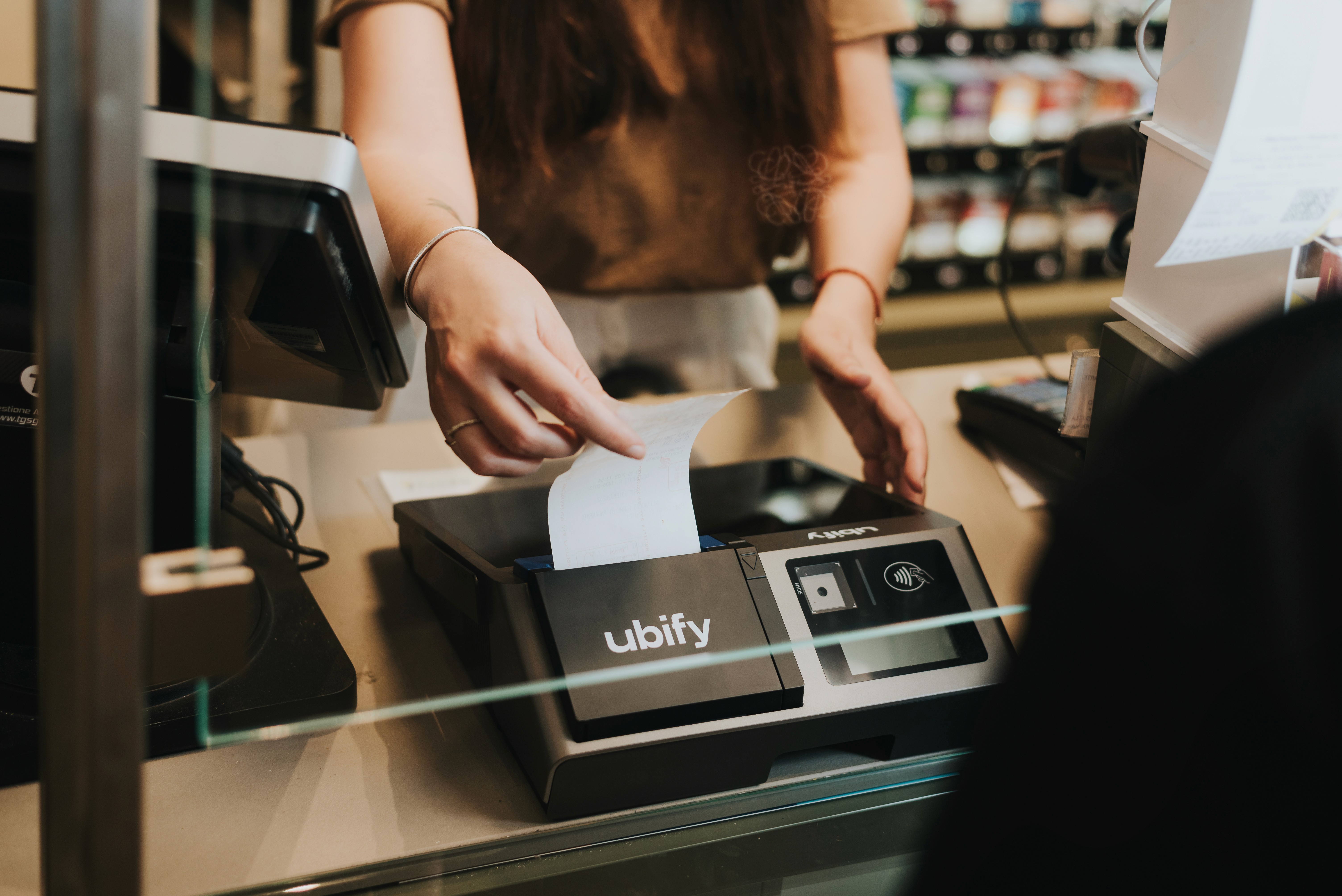 Commerciante consegna uno scontrino stampato con la Cassa Ubify, vista attraverso il vetro del bancone.