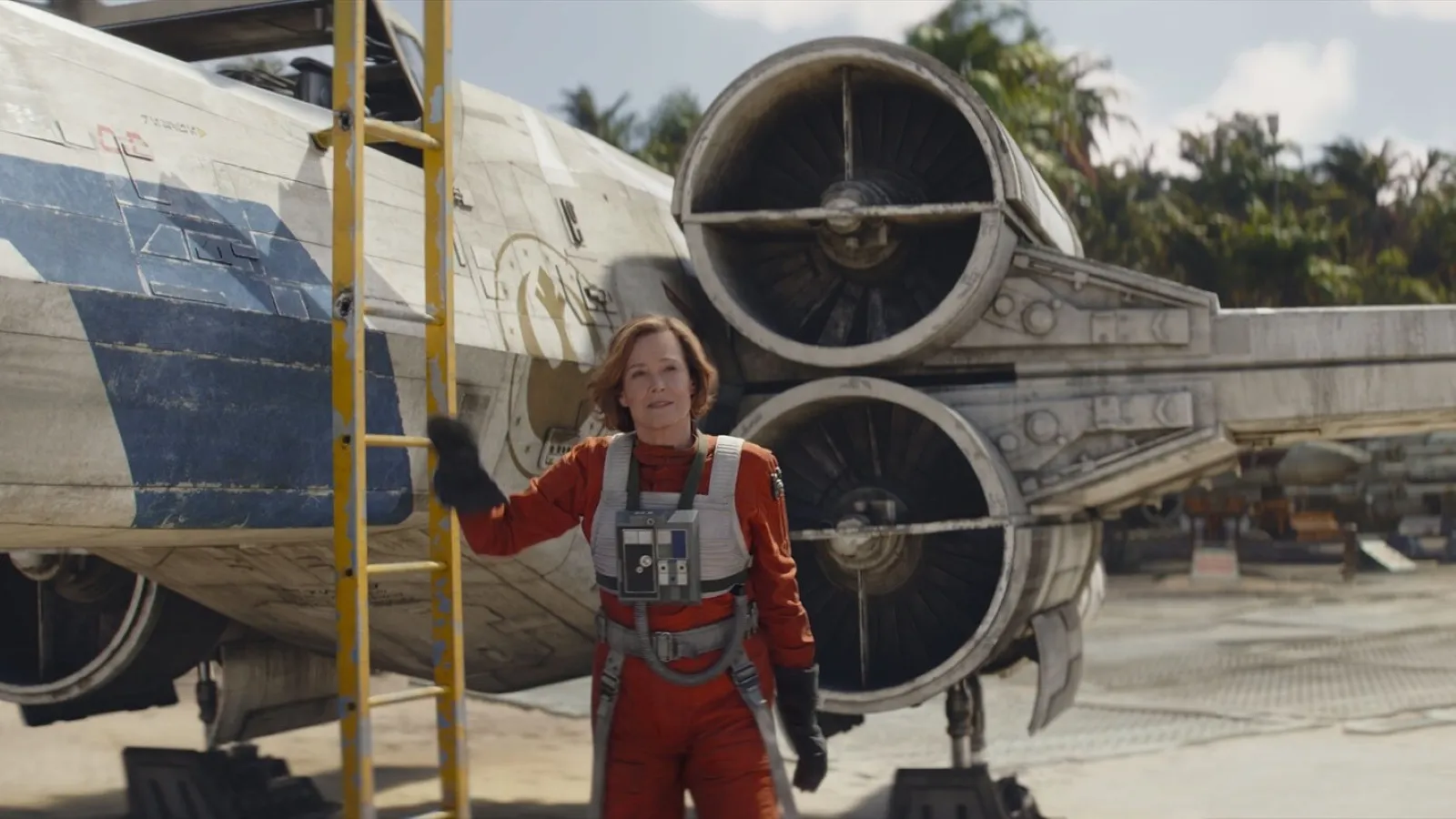 A woman in a red flight suit stands beside a large space fighter with circular engines, set against a backdrop of palm trees, suggesting a tropical location.