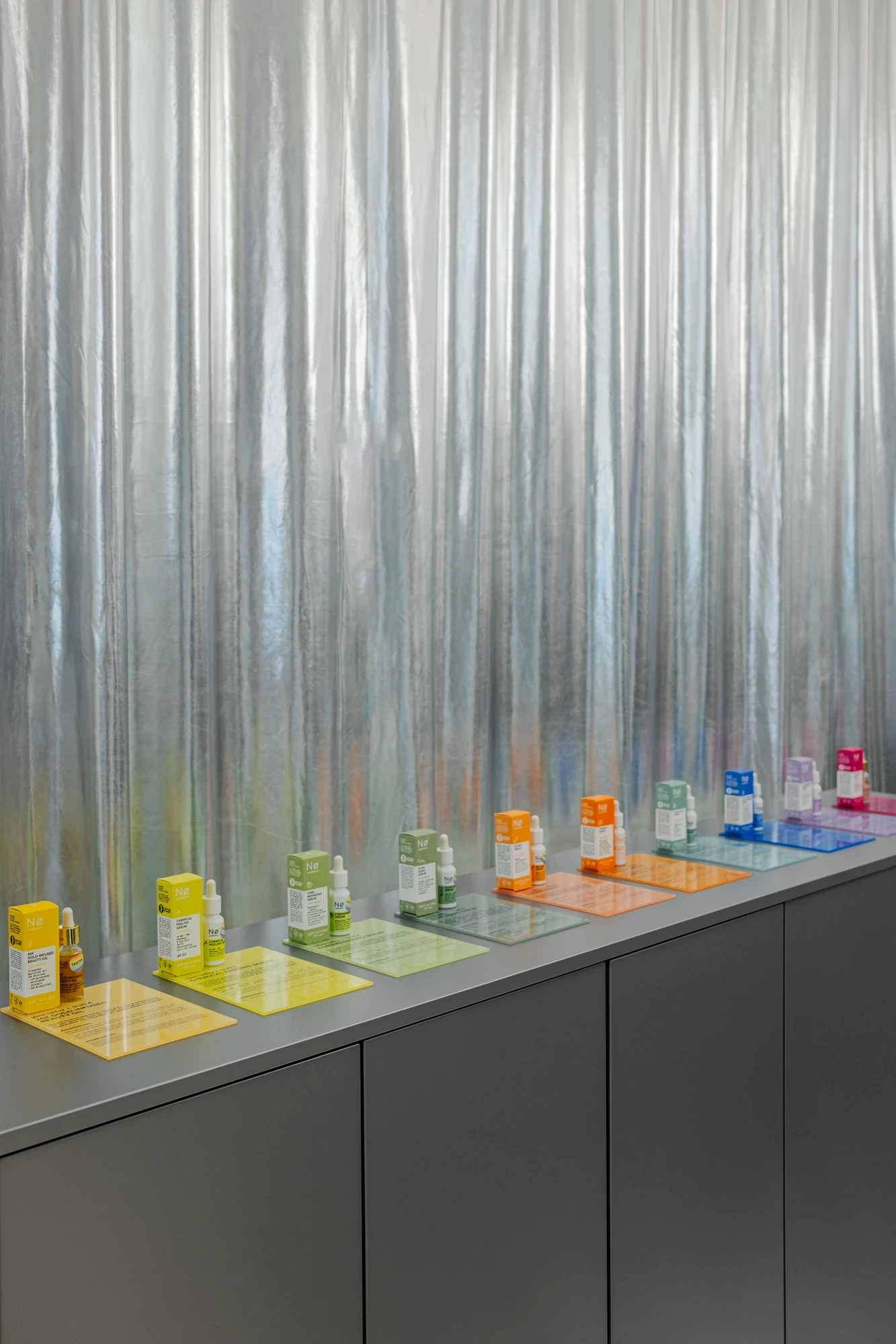 Product display lined up on a counter with colourful translucent cosmetics packaging in front of a metallic curtain backdrop.