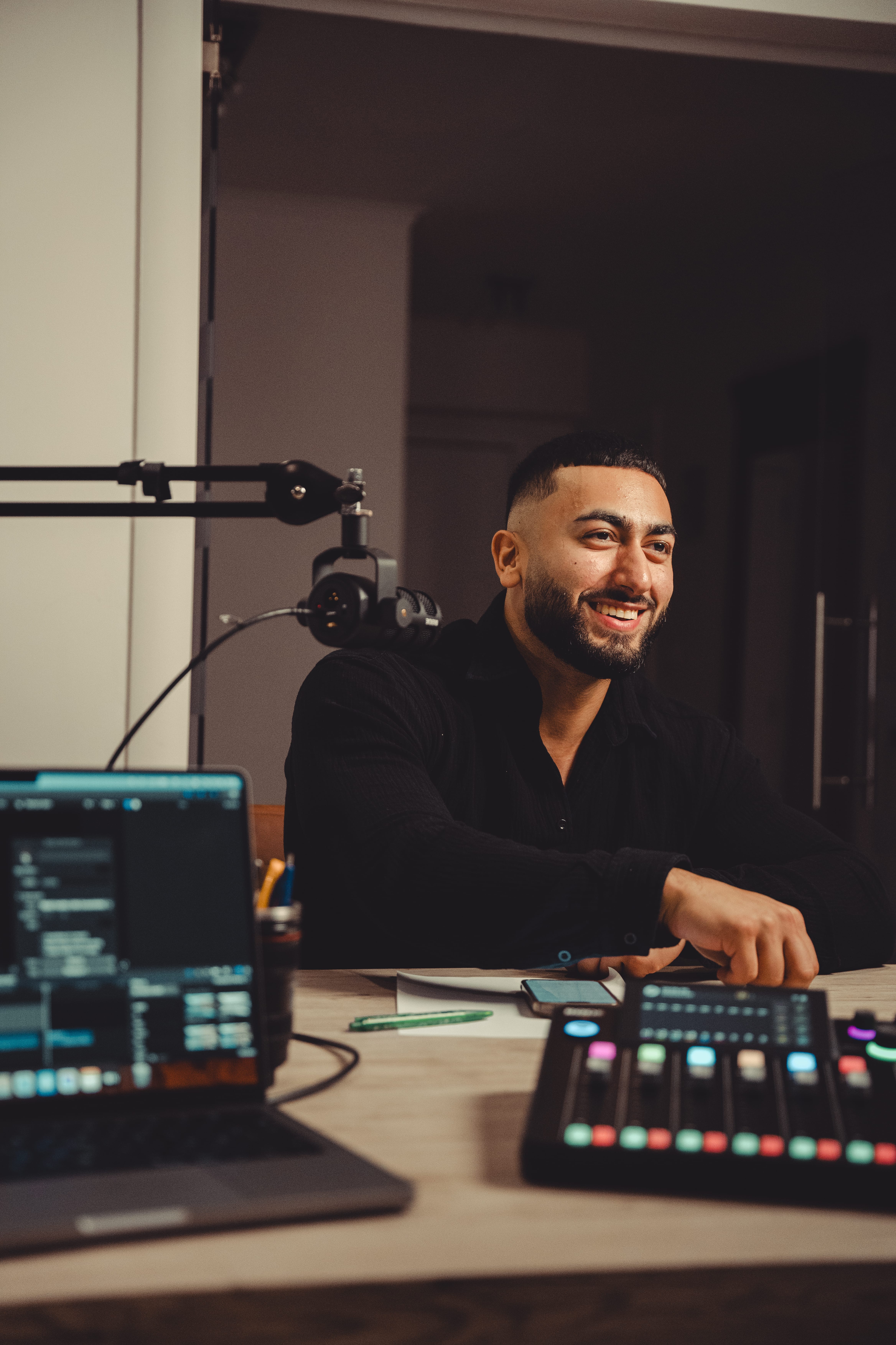 Sohil Elyas van Sohil Coaching kijkt lachend naar de interviewer terwijl hij op een stoel zit in een podcast-setting, met een opvallende microfoon in beeld.