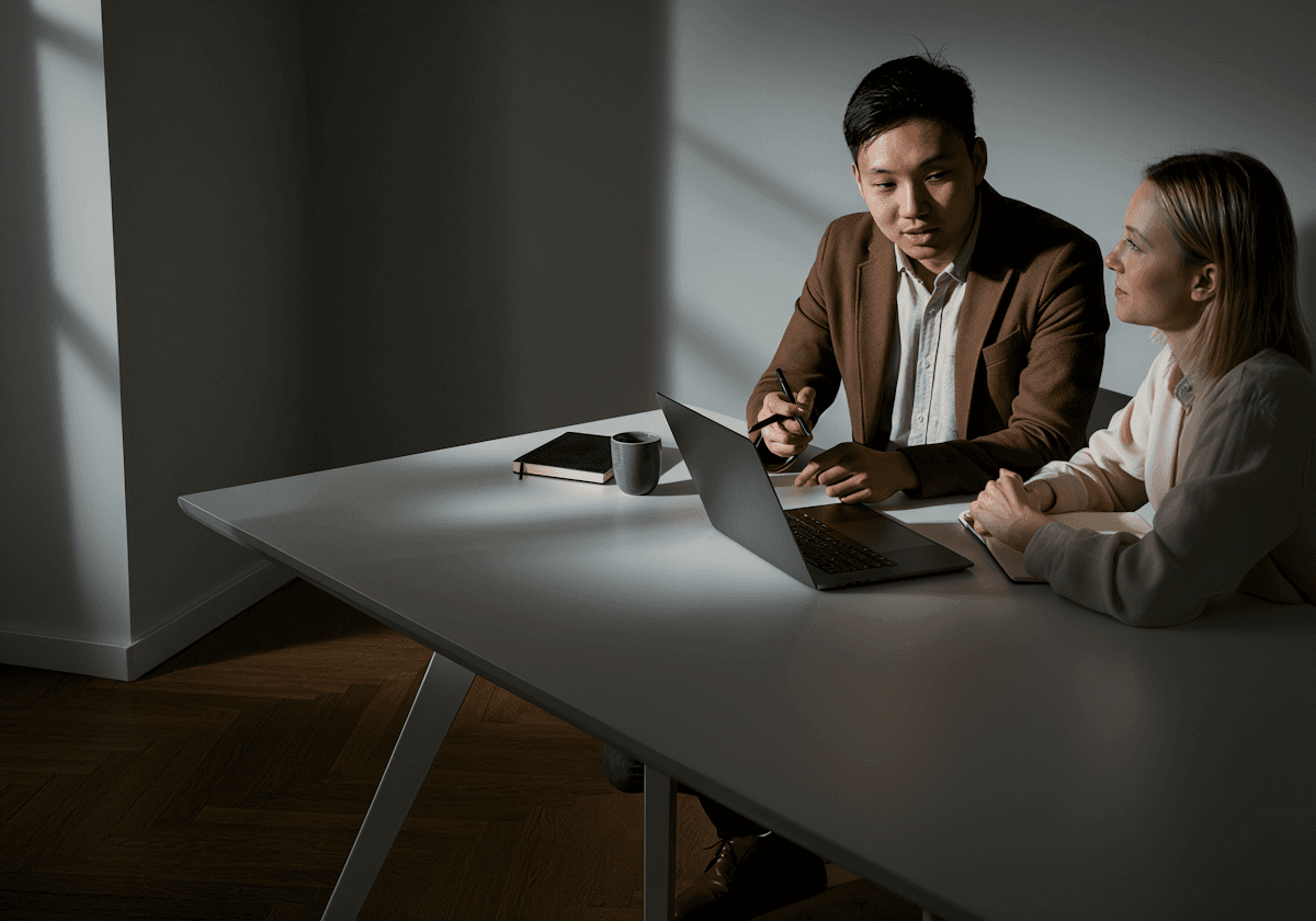 Two people sit at a white table in a dimly lit room, discussing something. The man gestures towards a laptop, while the woman listens attentively.