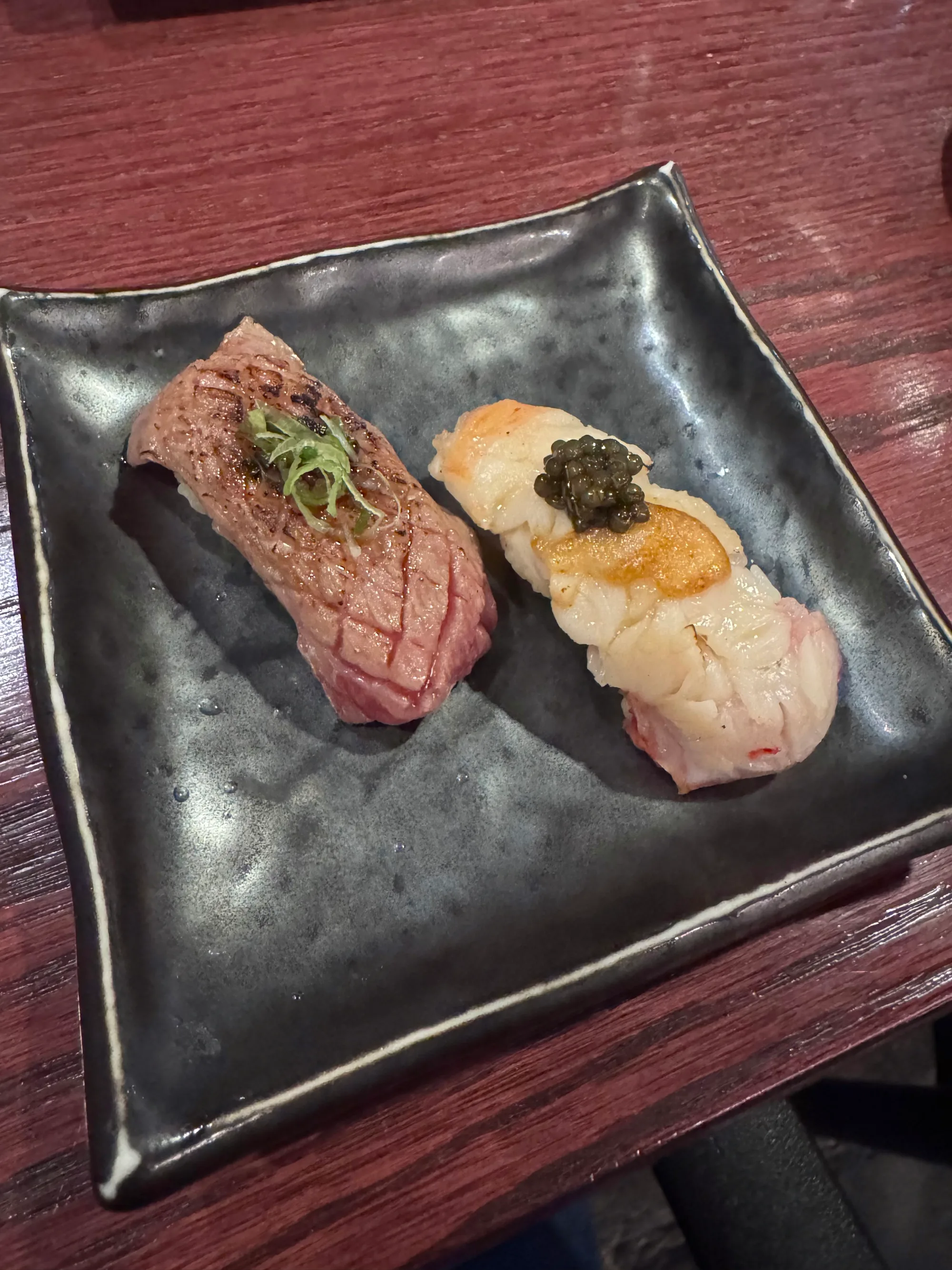 Two pieces of gourmet sushi, Wagyu beef nigiri and scallop with caviar, on a black plate.