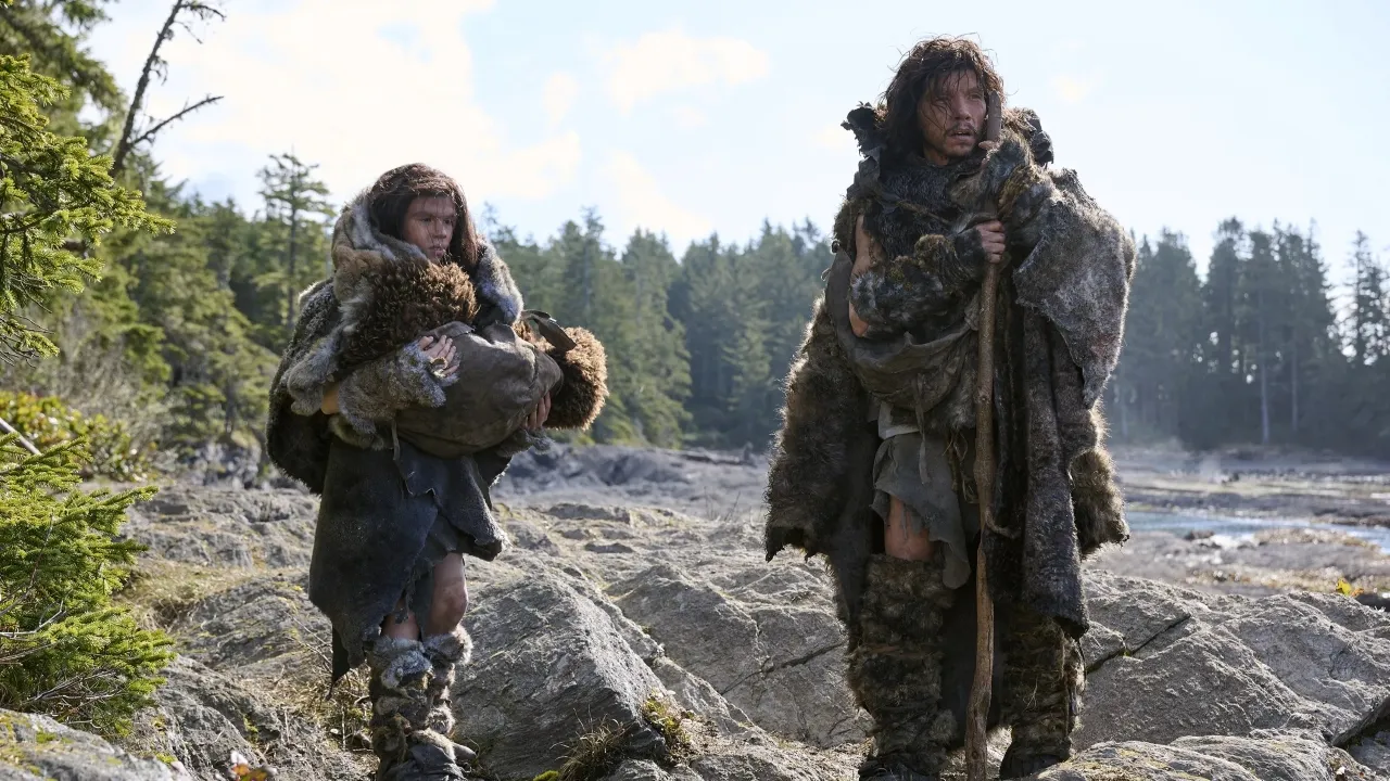 Two people wearing heavy fur clothing stand on rocky terrain near a body of water, surrounded by dense evergreen trees, portraying a prehistoric setting.