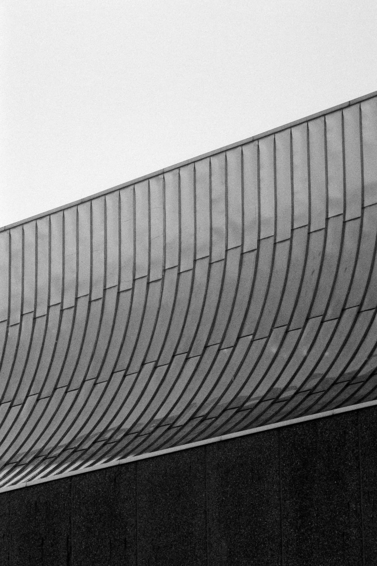 Curved corrugated metal cladding against blank sky, repeating vertical lines creating rhythmic pattern across diagonal architectural form