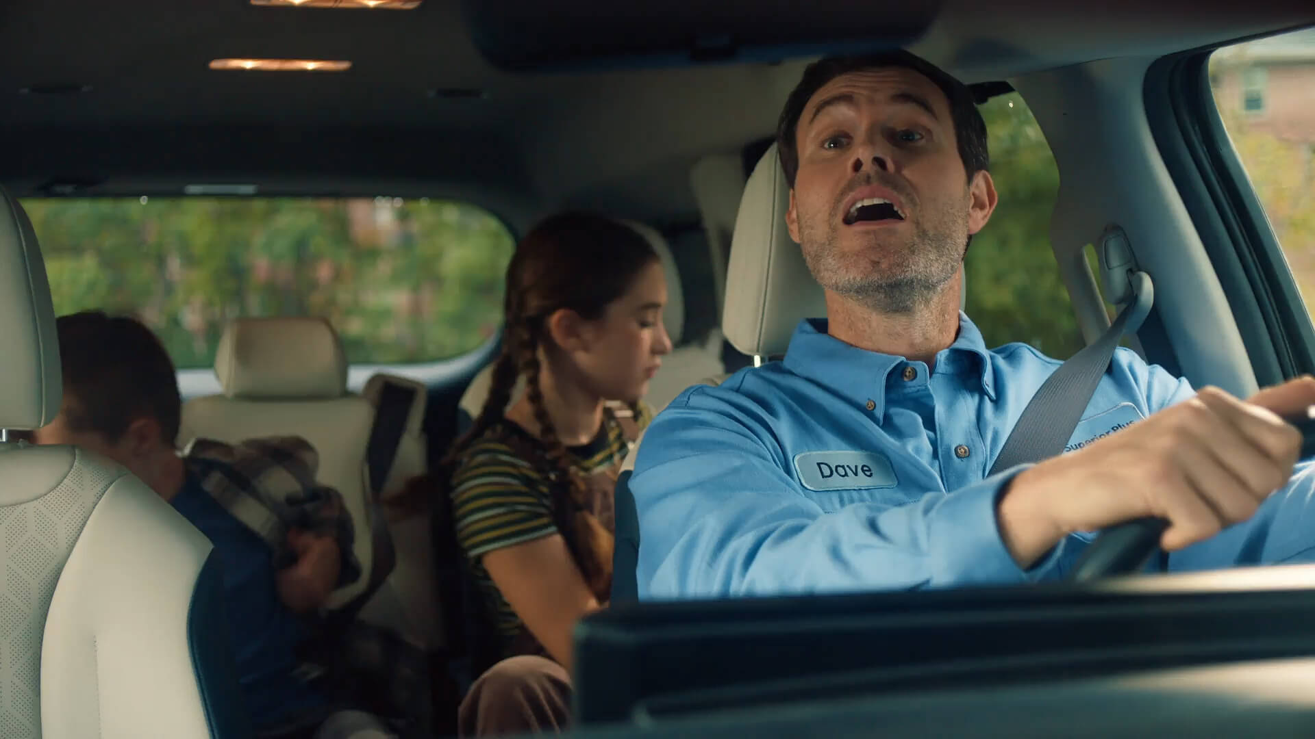 A man in a blue work shirt labeled "Dave" drives a car with two children sitting in the back seat.