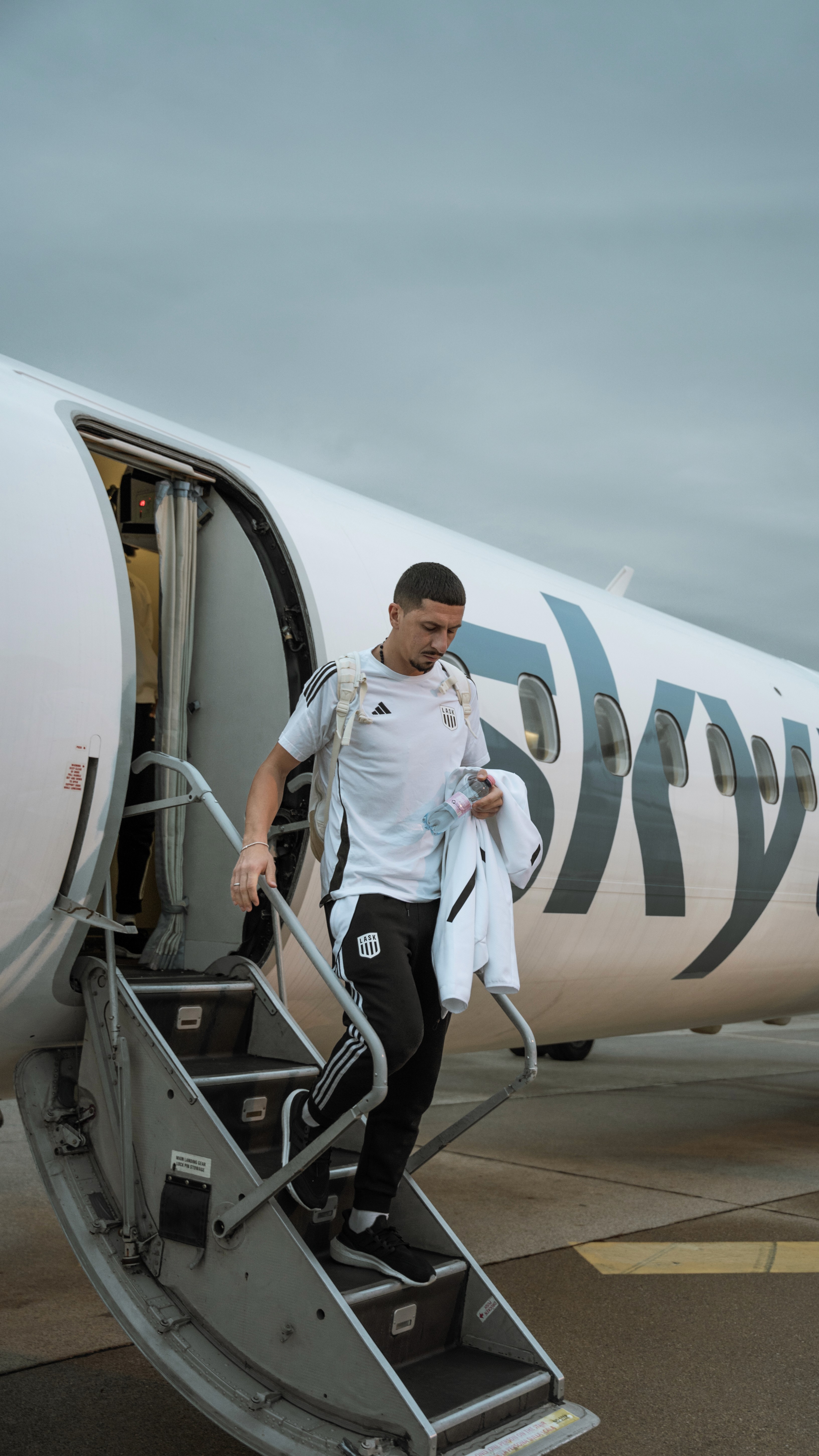 Fußballspieler steigt aus einem Flugzeug mit Sky-Logo. Er trägt Sportkleidung und einen Rucksack.
