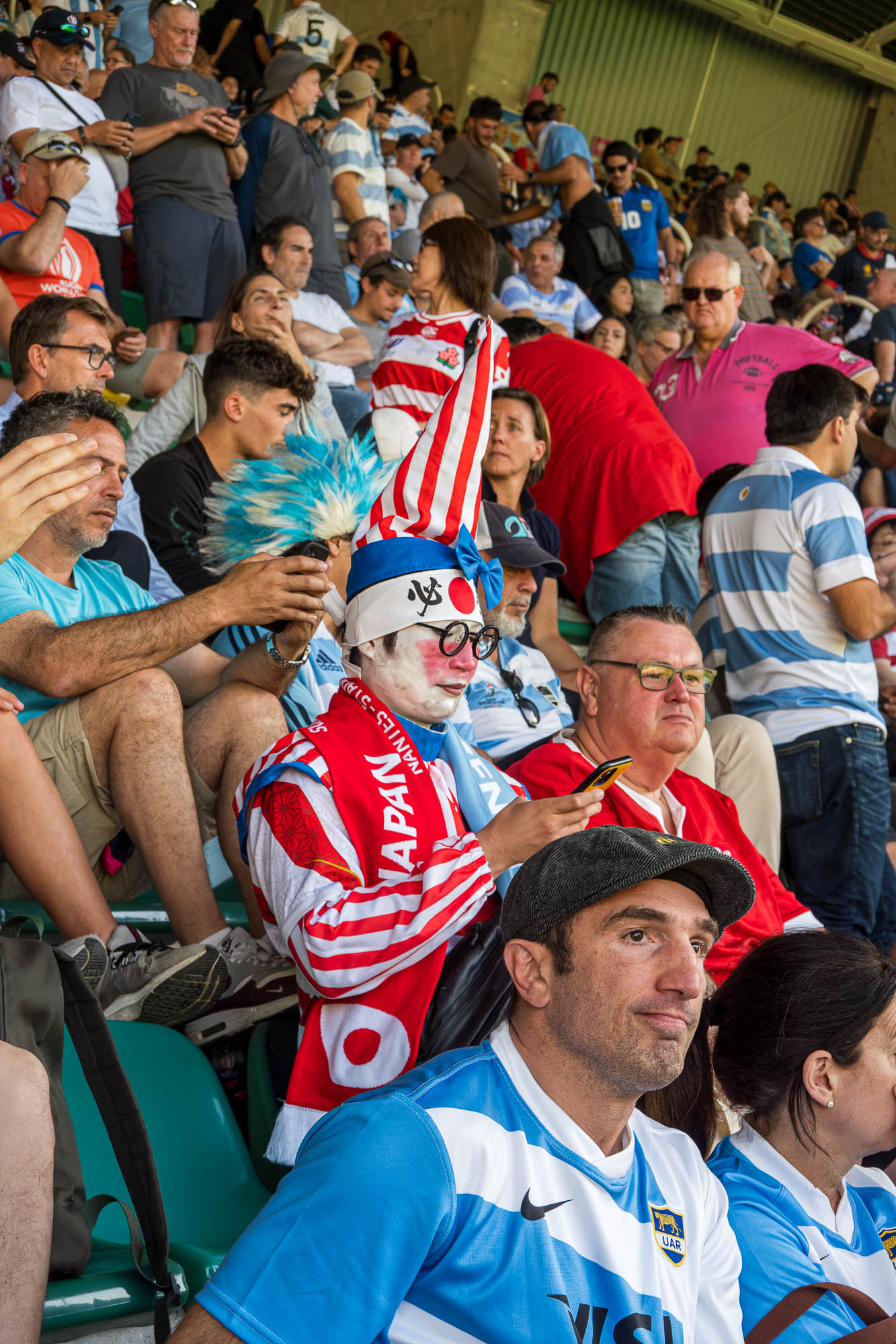 Supporter japonais au bonnet pointu rayé rouge et blanc, visage poudré et lunettes rondes, consultant son téléphone derrière un supporter argentin en maillot ciel et blanc