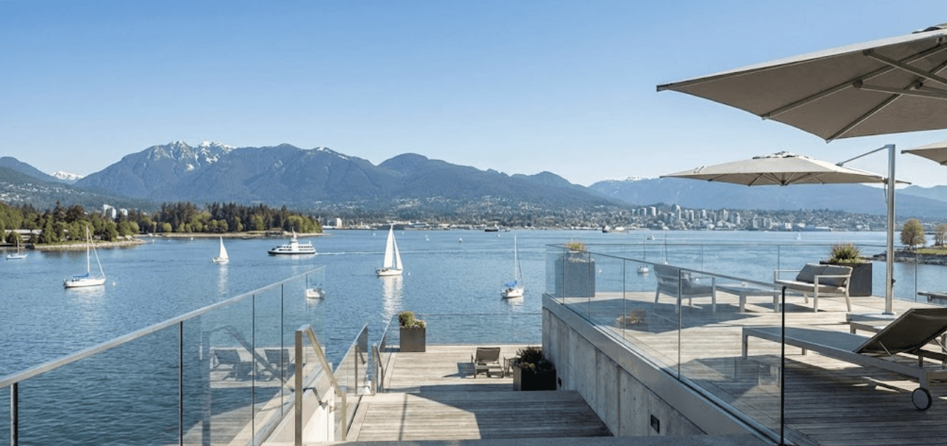 Modern waterfront deck with frameless glass railings overlooking Vancouver harbor, clear blue sky, sailboats in the distance, North Shore mountains in background, professional architectural photography