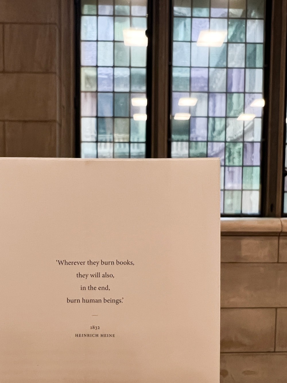 De eerste pagina van het geopende boek met een centrale quote. Op de achtergrond zijn de karakteristieke glas-in-loodramen van de Universiteitsbibliotheek van de KU Leuven zichtbaar, wat de historische context van het project benadrukt.