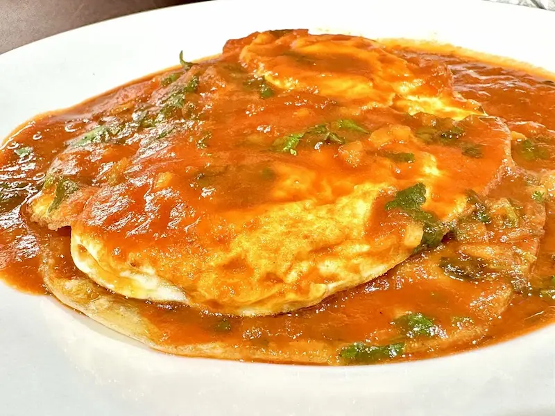 A vibrant plate of huevos rancheros with sunny-side-up eggs smothered in red ranchero sauce, garnished with fresh cilantro, served on a white dish at Oneco Farmers Market, Bradenton, FL.