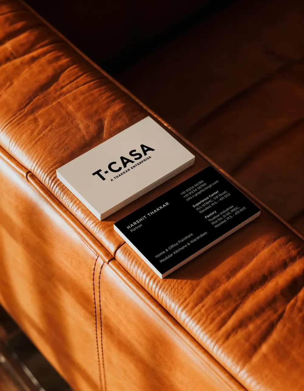 Set against a rich, brown leather sofa, a stack of white and black business cards is presented in a warm, sunlit environment, showcasing sleek typography and a minimalist design.