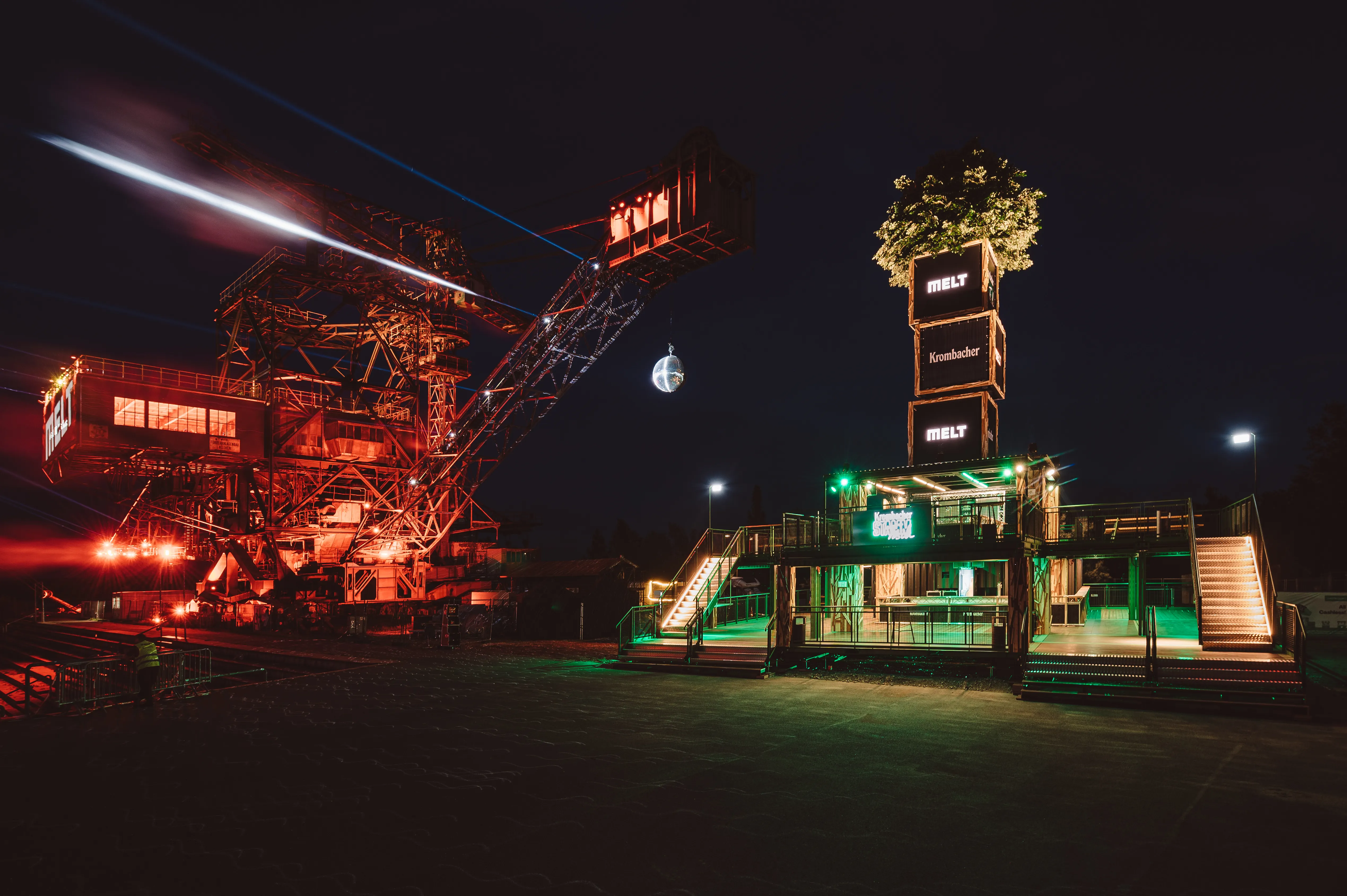 Night view of the KROMBACHER tower with glowing Melt and Krombacher logos beside the red-lit excavator cranes of Ferropolis.