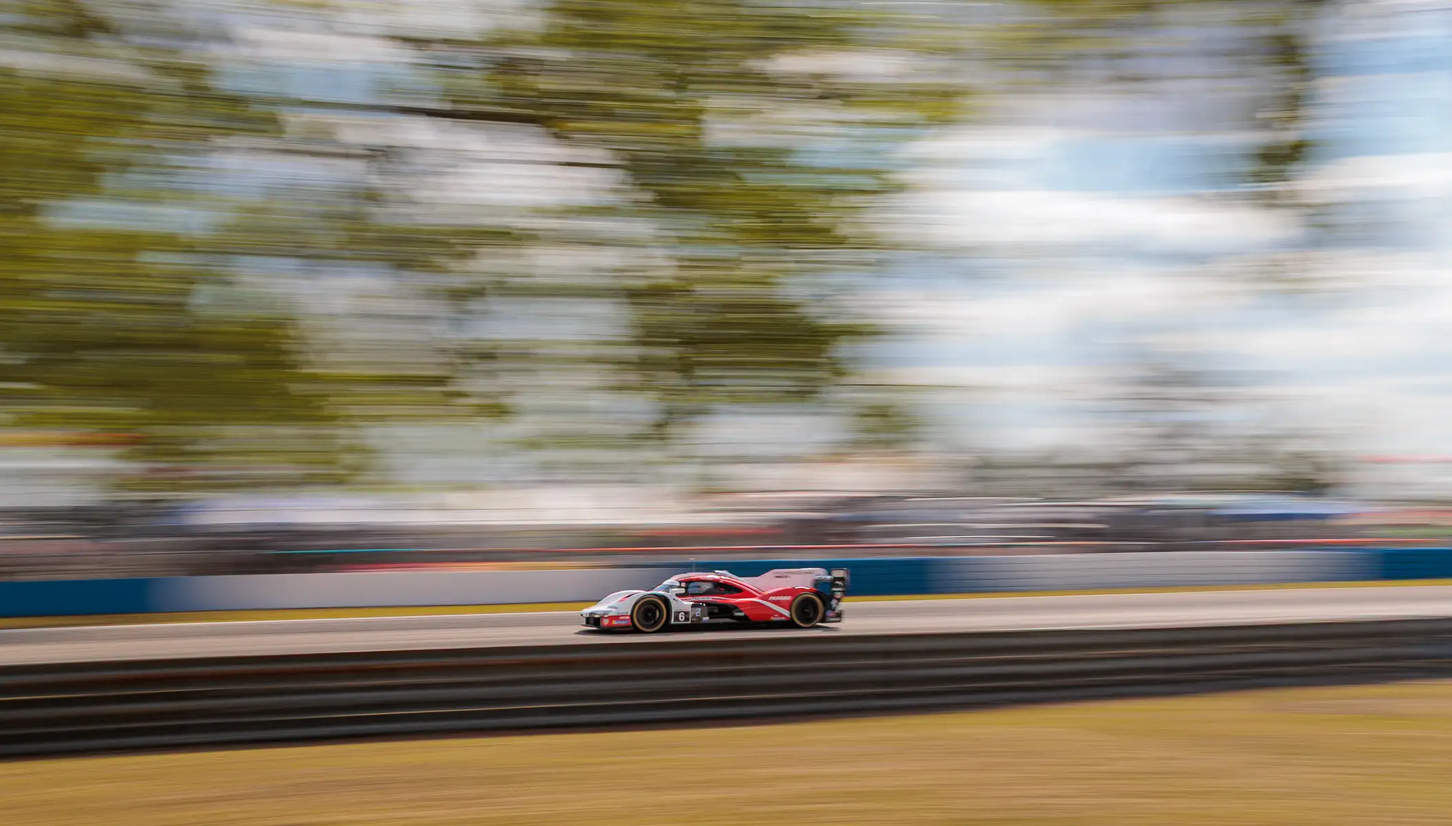 A red Porsche racing car speeds down a track, against a blue sky and trees, with a motion blurred background.