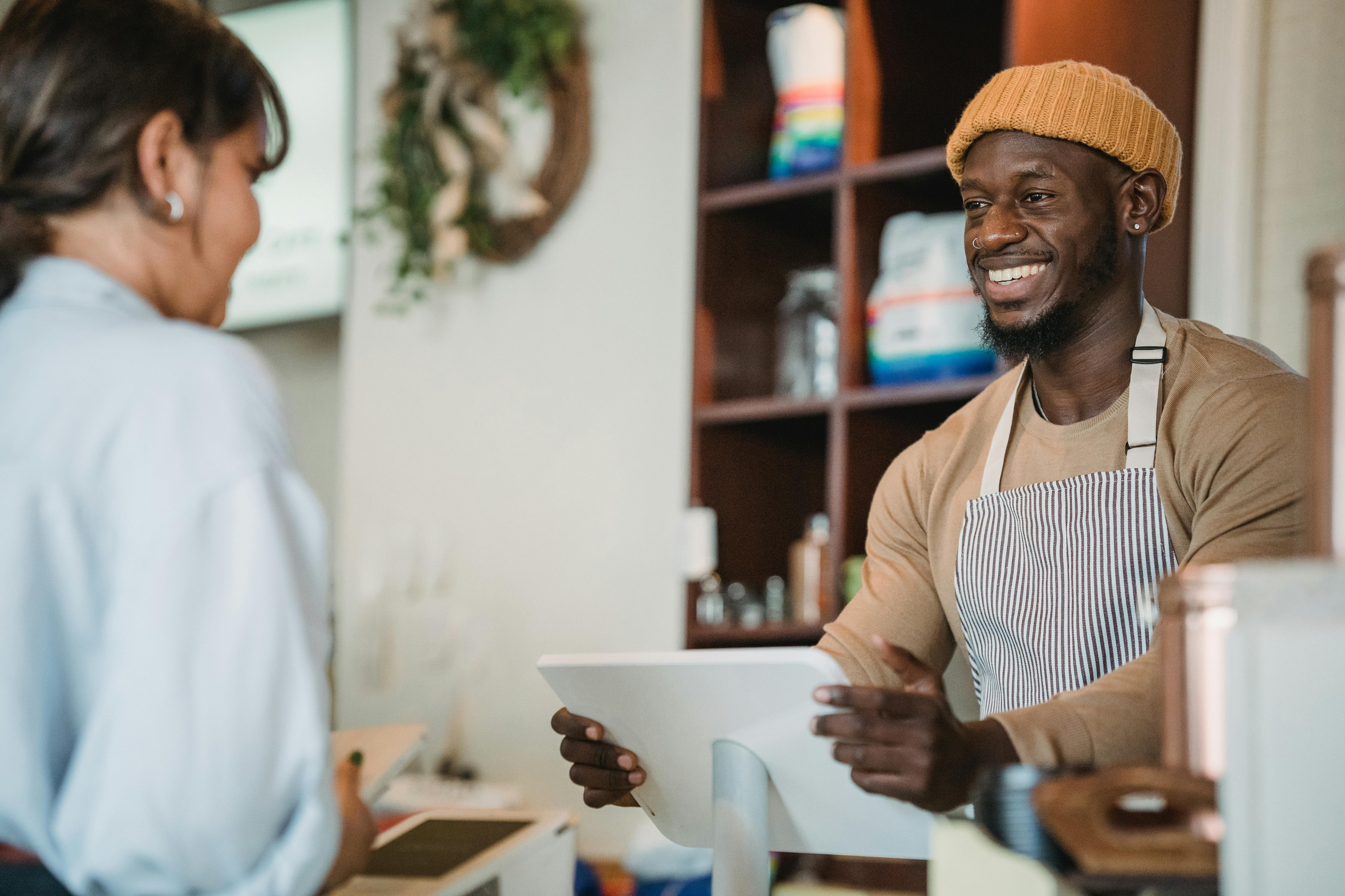 barista-with-apron-and-beanie-smiling-as-he-takes-an-order-from-a-customer-in-a- - mizuno-k (pexels)