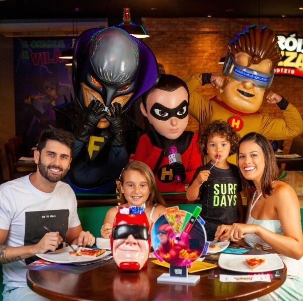Família sentada à mesa ao lado de personagens fantasiados em pizzaria temática voltada ao público infantil.
