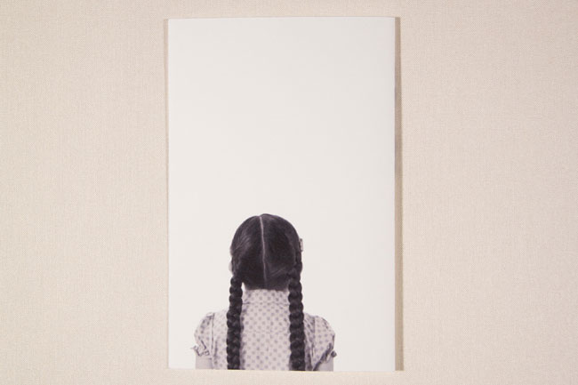 A person with long black braids stands with their back to the camera, wearing a light-colored shirt with small polka dots. The image is framed by a white border against a light beige wall. The person’s hair is centered in the frame, and the image is centered on the white border.