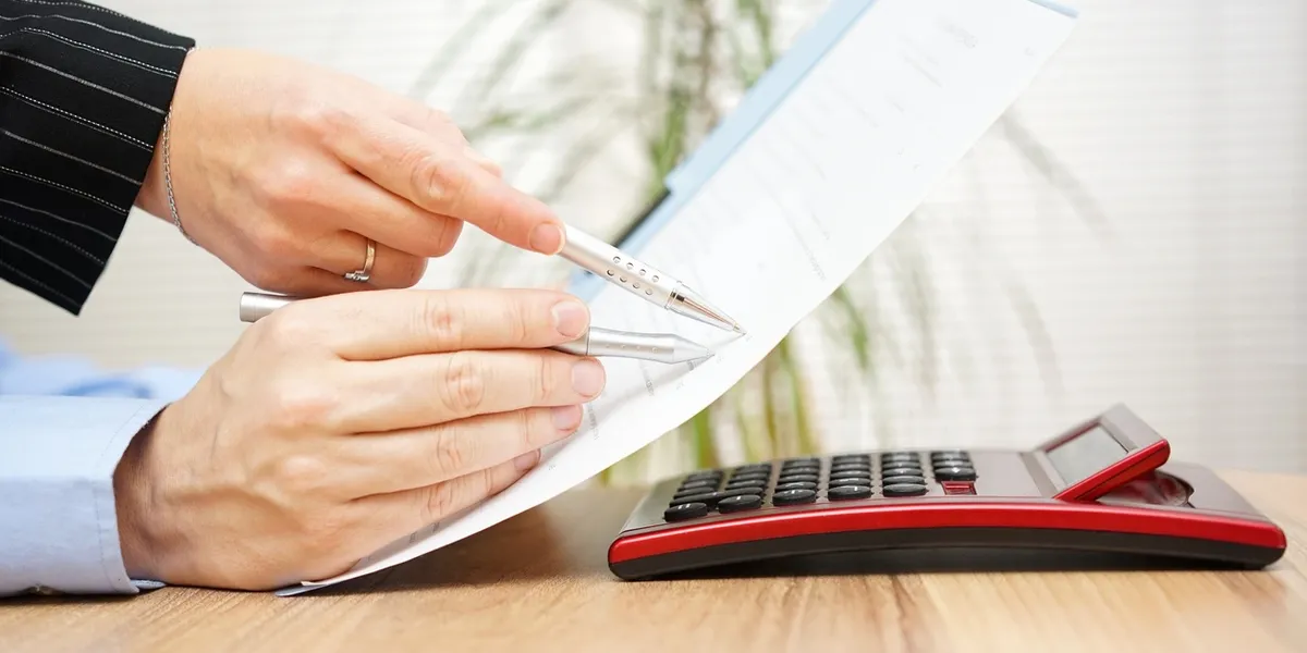 A close-up of two people pointing pens at a printed document on a desk next to a red calculator.