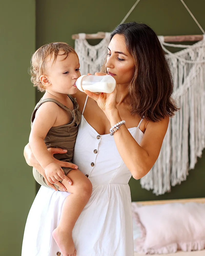 A mother in a white dress holding and bottle-feeding her toddler in a boho-styled nursery