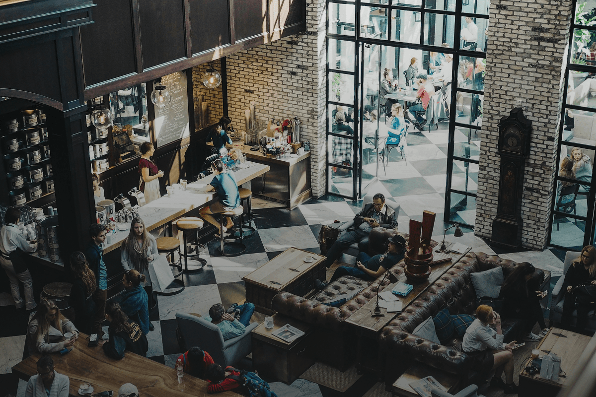 Bird’s-eye view of a lively coffee shop with customers enjoying coffee. Some are browsing on their phones, others are working on laptops or chatting. A barista is preparing drinks at the coffee bar.