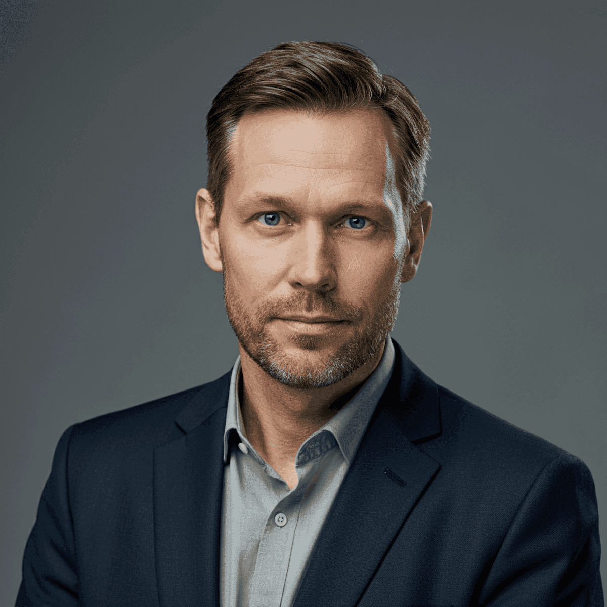 Professional headshot of a man with short brown hair and a light beard. He wears a navy blazer and gray shirt, conveying confidence and approachability.