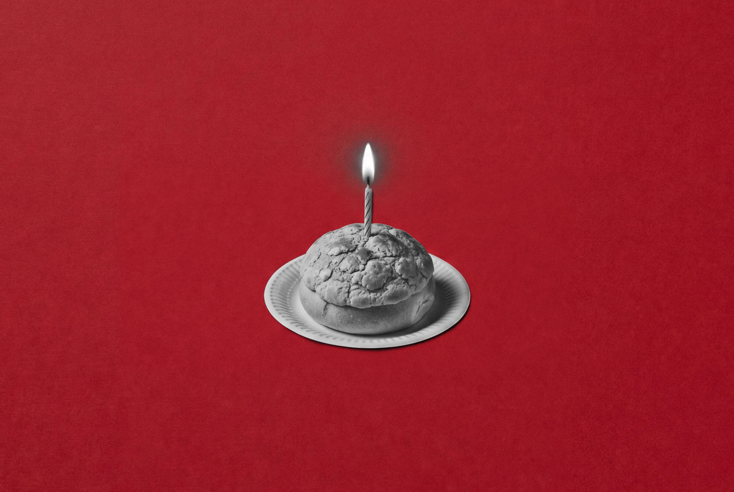 A black-and-white pineapple bun on a paper plate with a single lit birthday candle stuck in the top, on red paper.