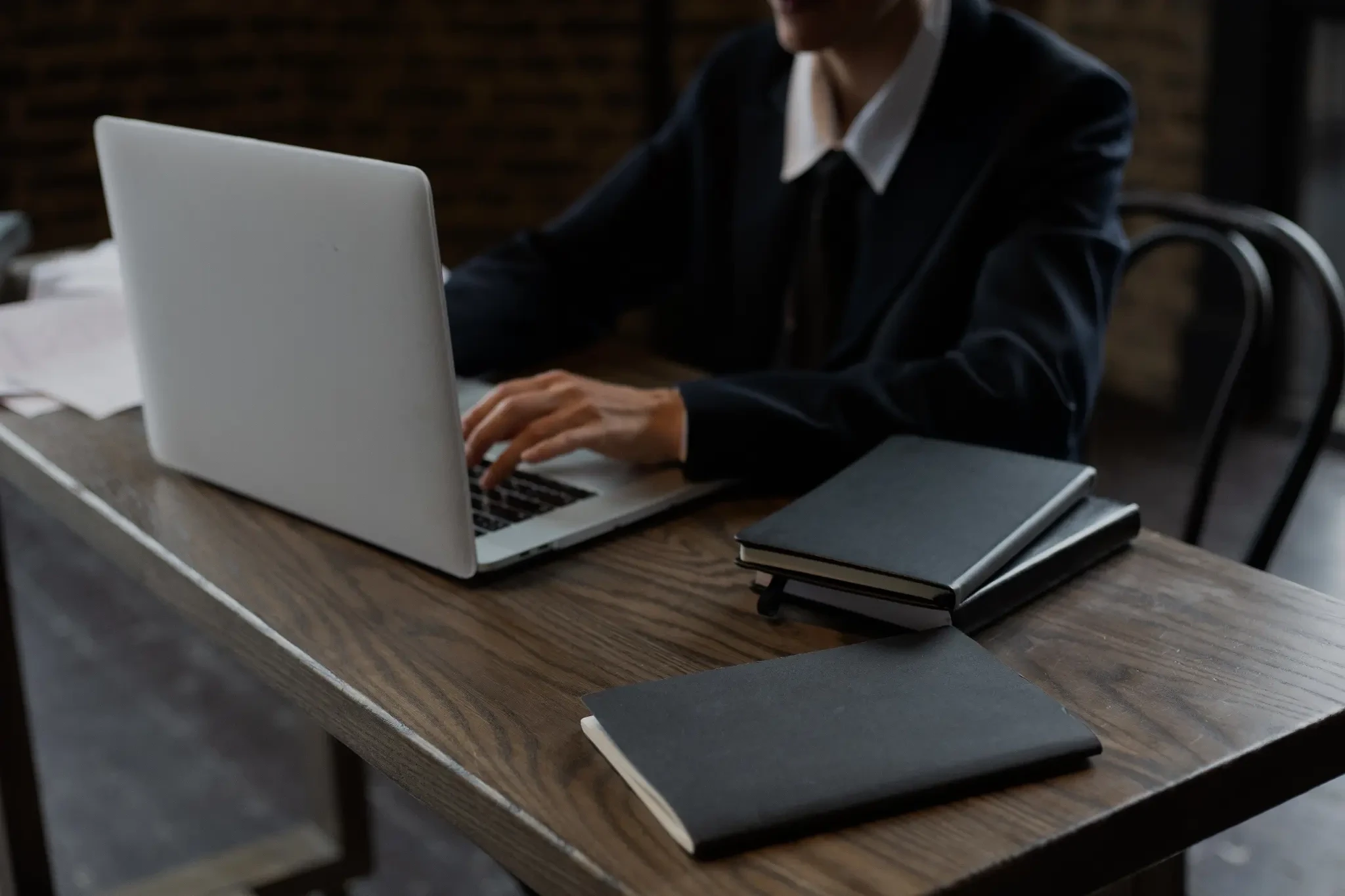 Professional working on a laptop at a wooden desk with notebooks.