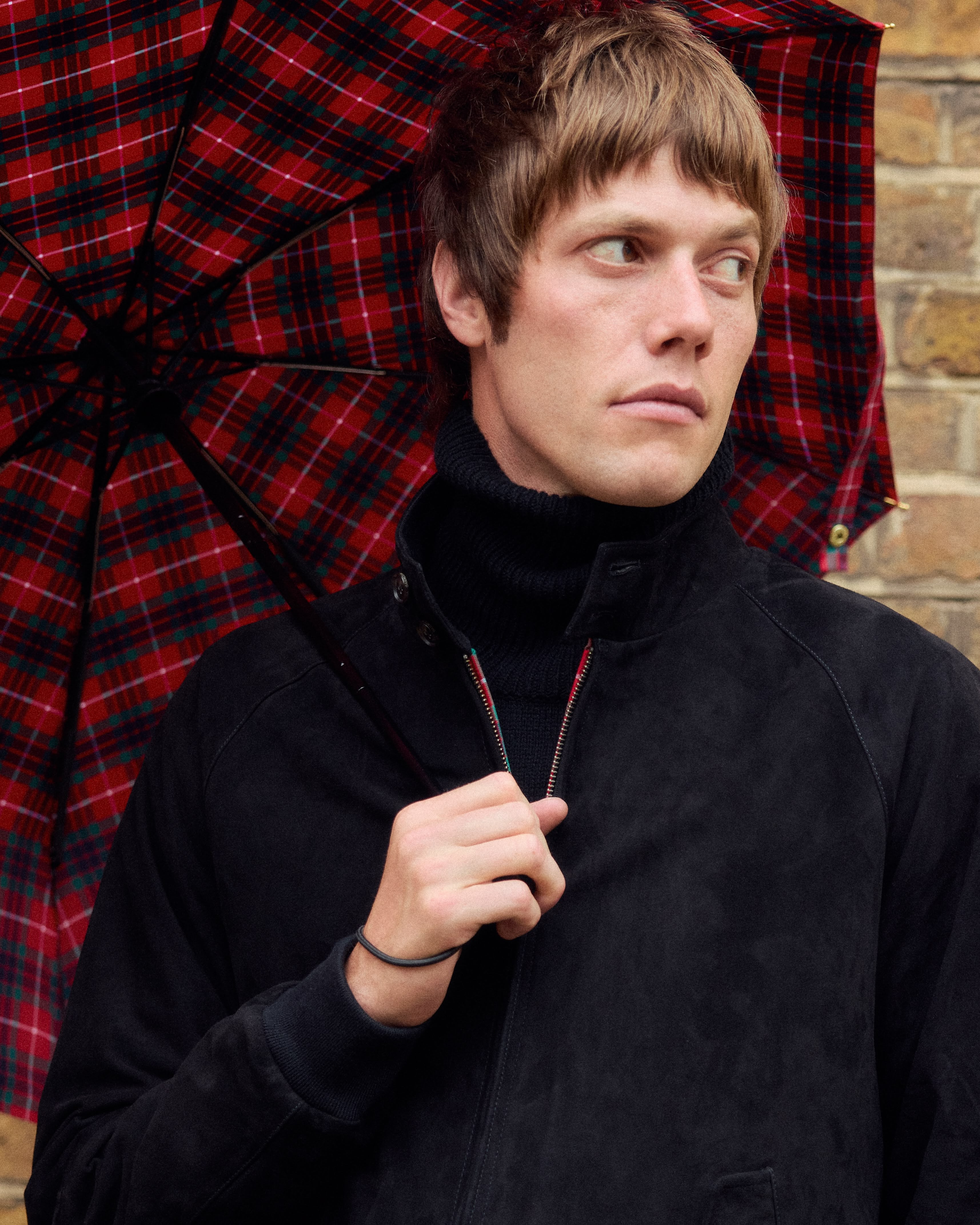 Le mannequin avec une coupe Beatles porte blouson noir sur col roulé noir avec un parapluie tartan de la marque Baracuta