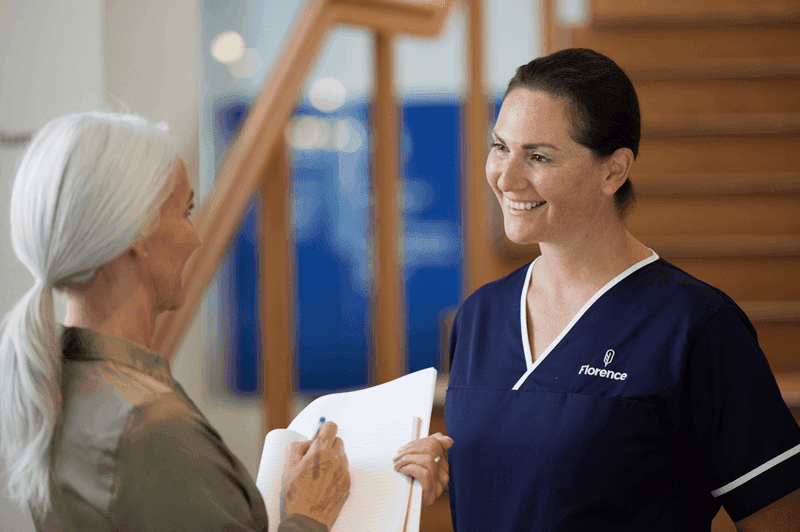 Florence caregiver in uniform smiling while speaking with an older woman holding documents indoors.