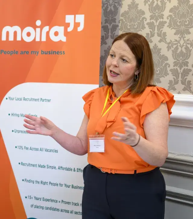 A woman in an orange blouse gestures while speaking, standing in front of an orange recruitment banner. The setting conveys a professional tone.