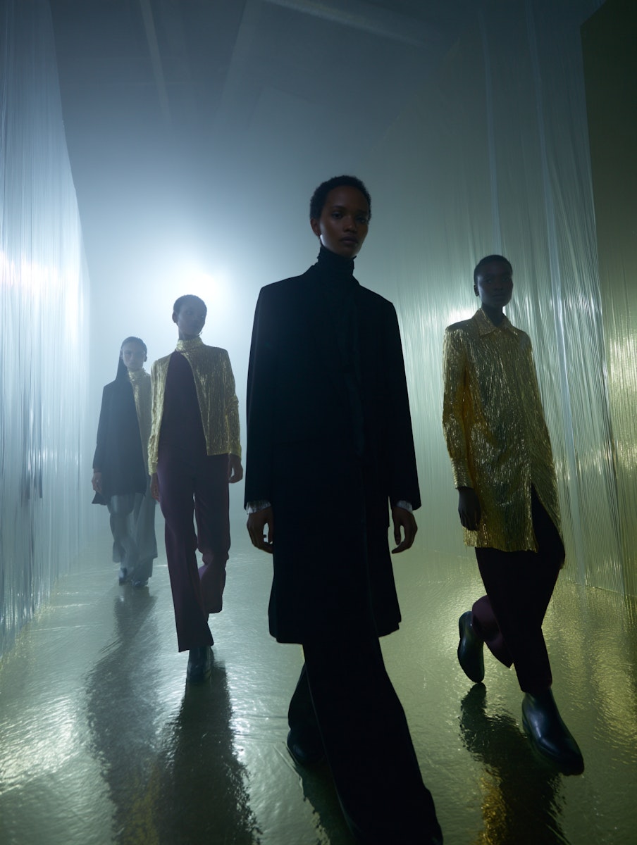 Four models walk a reflective runway in futuristic gold and black outfits, surrounded by ethereal lighting and translucent curtains.