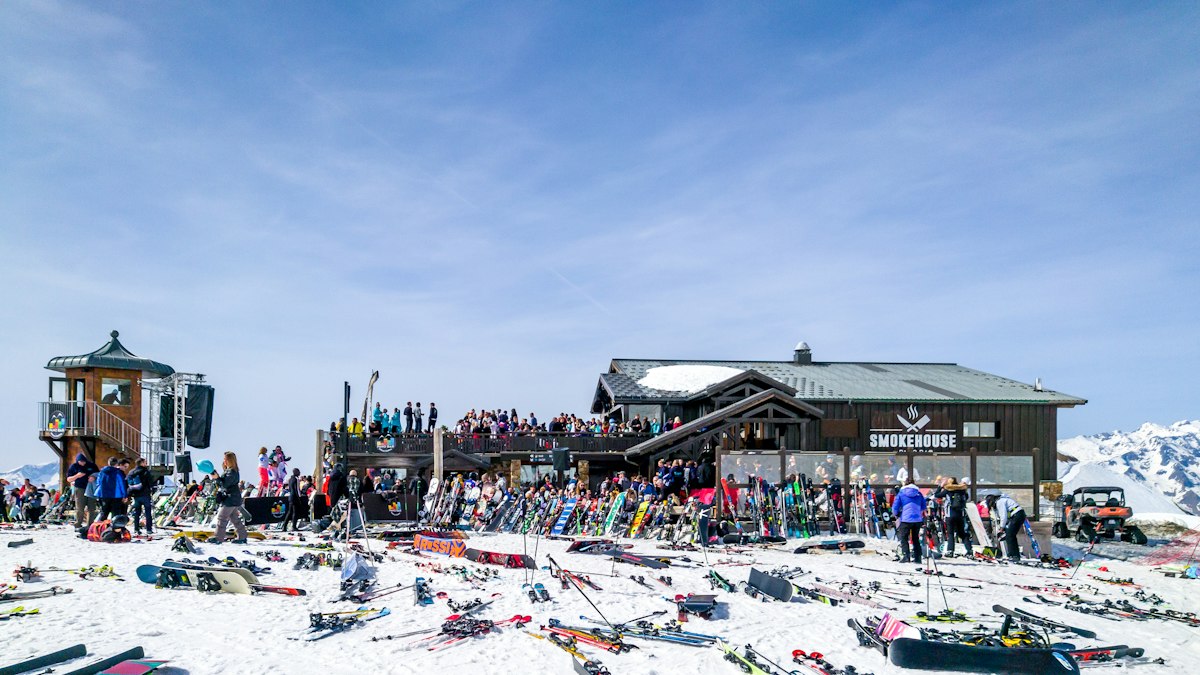 Hundreds of pairs of skis stacked in the snow outside a crowded mountain bar on a bright day