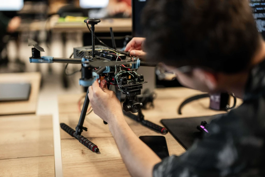A person is adjusting the components of a quadcopter drone, which is placed on a wooden desk in a tech workspace, surrounded by various electronic equipment and blurred monitors in the background.